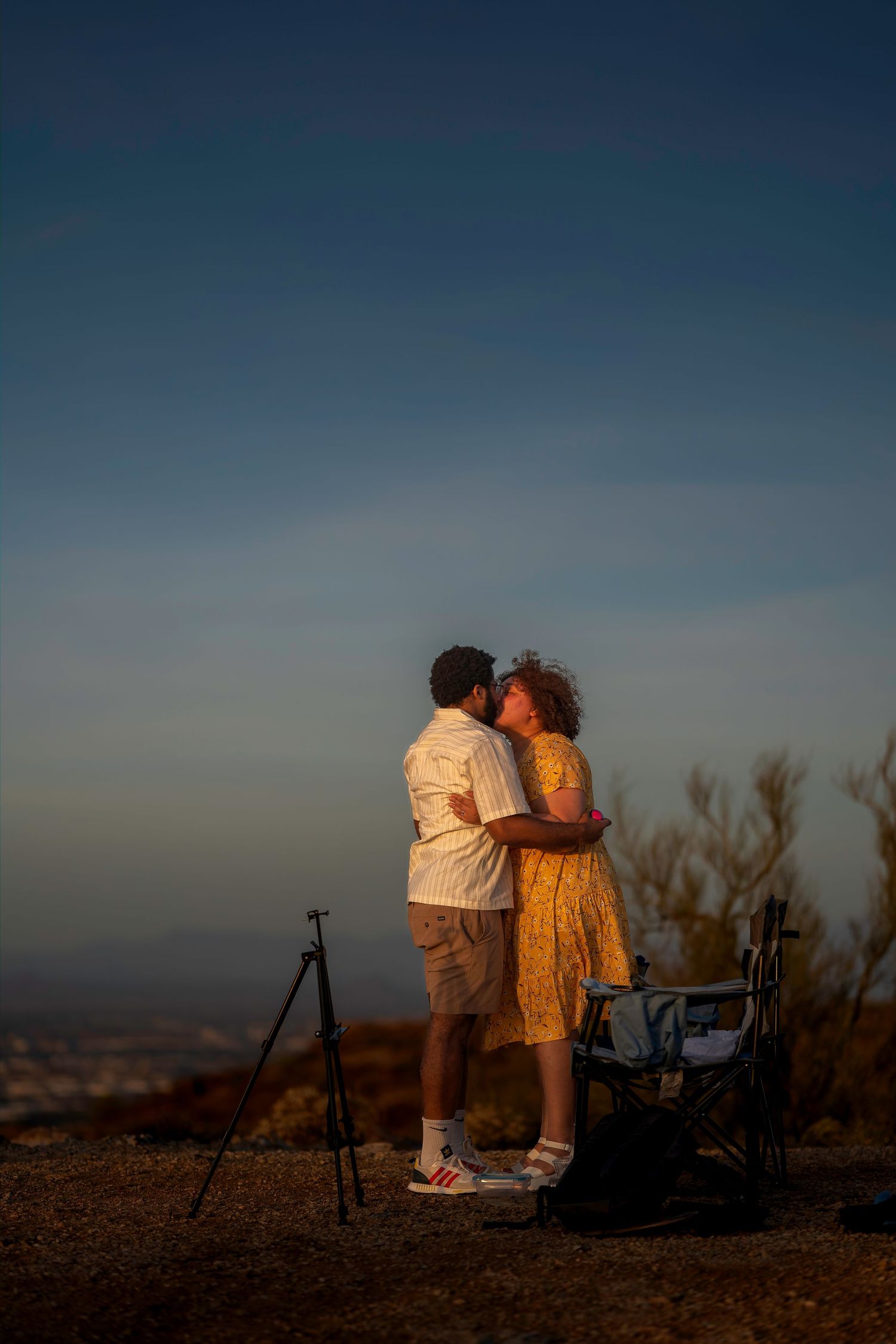 Couple sharing a romantic kiss at sunset on a hilltop with a tripod and city views in the background.