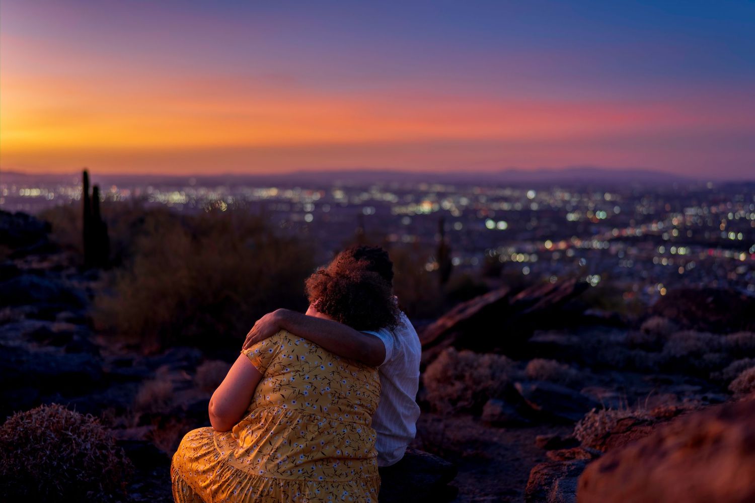 Couple hugging while watching a colorful sunset over a glowing city skyline.