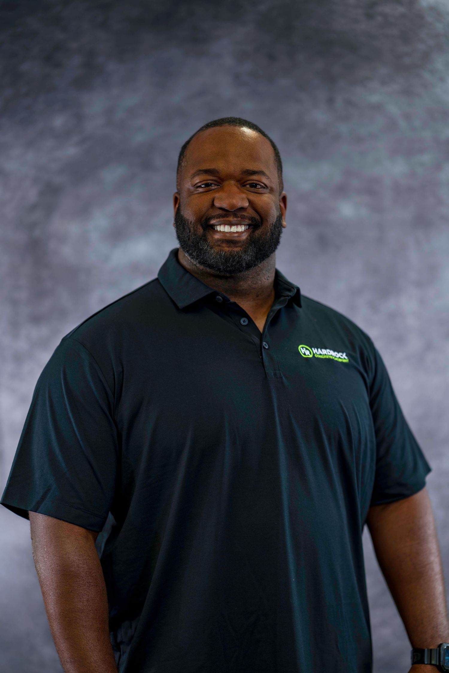 Professional headshot of bearded employee in dark polo shirt against textured gray background.
