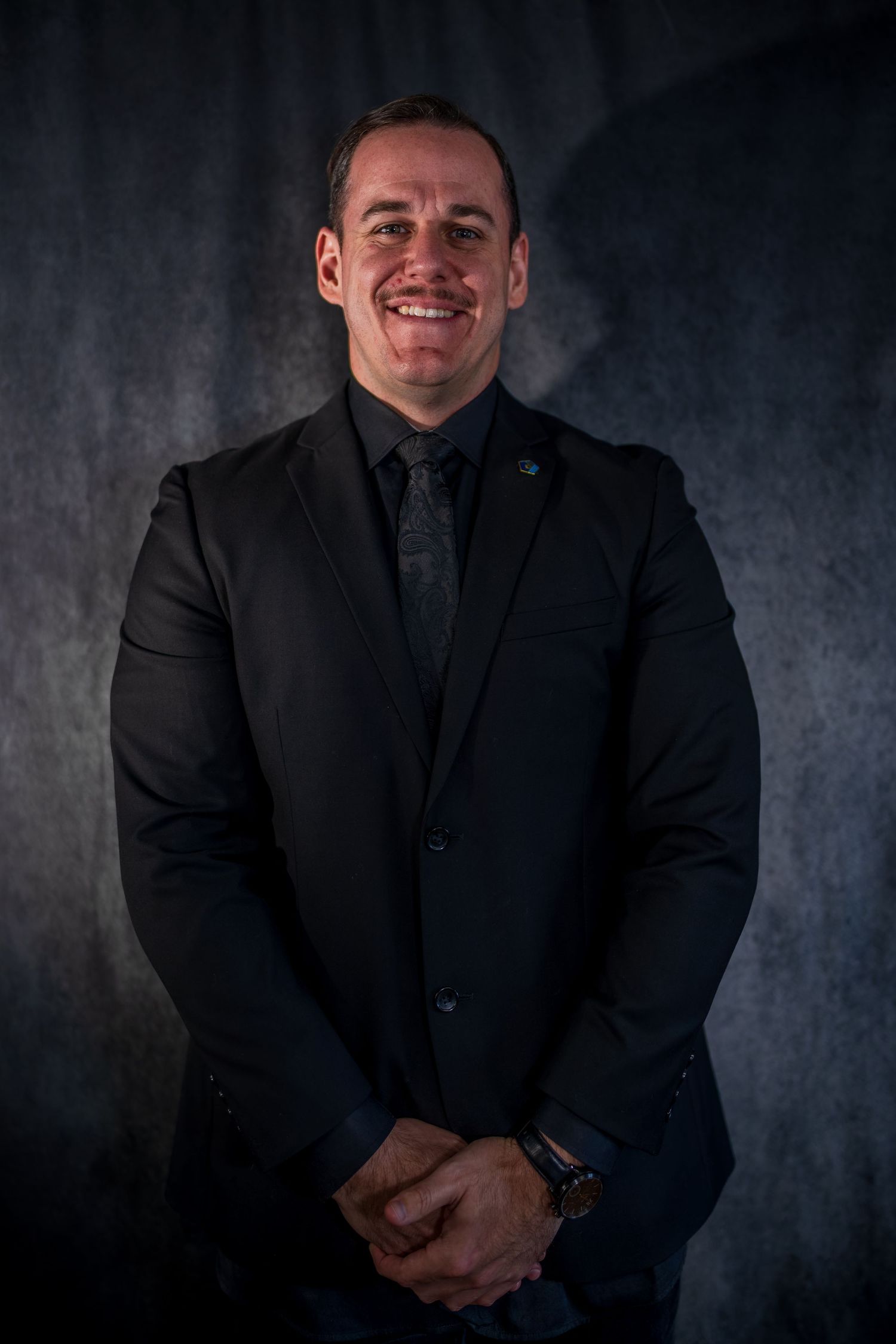 Professional portrait in all-black attire against dramatic dark studio background.