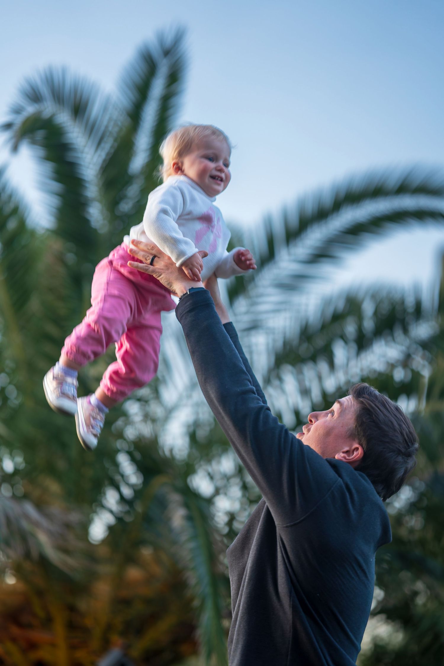 Parent lifting and playing with a baby wearing pink clothes against a palm tree background.