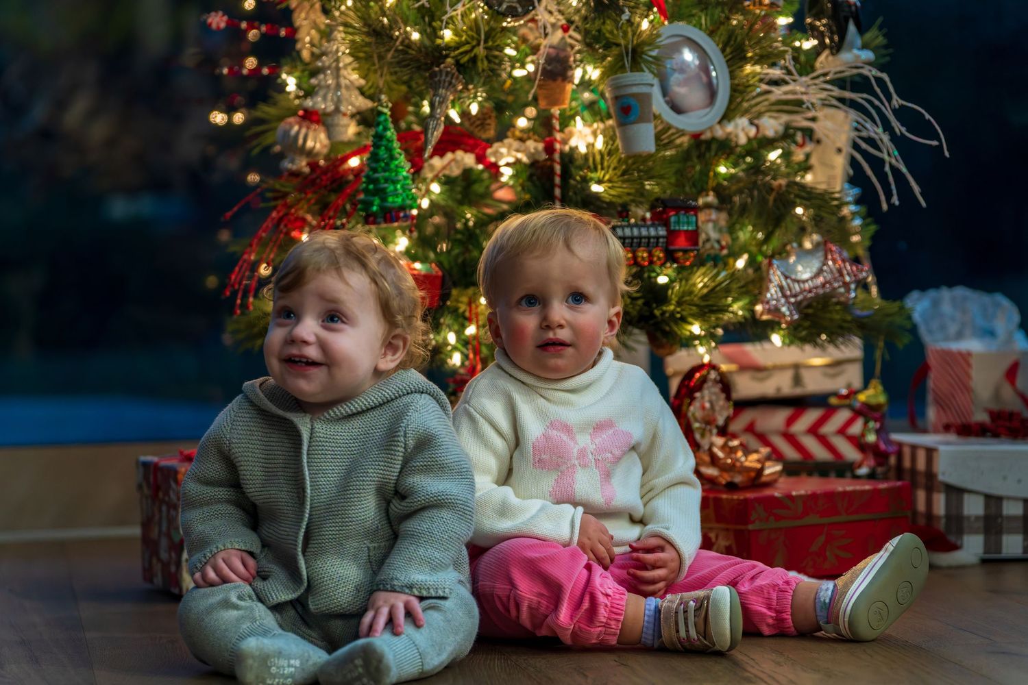 Two toddlers sit together in front of a decorated Christmas tree with presents.