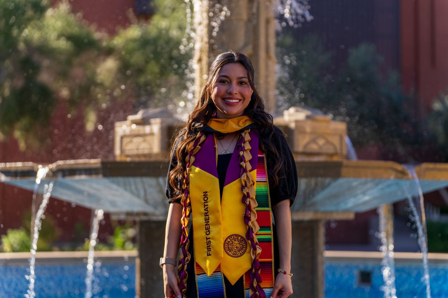 Graduate in colorful stole stands near campus fountain on graduation day.