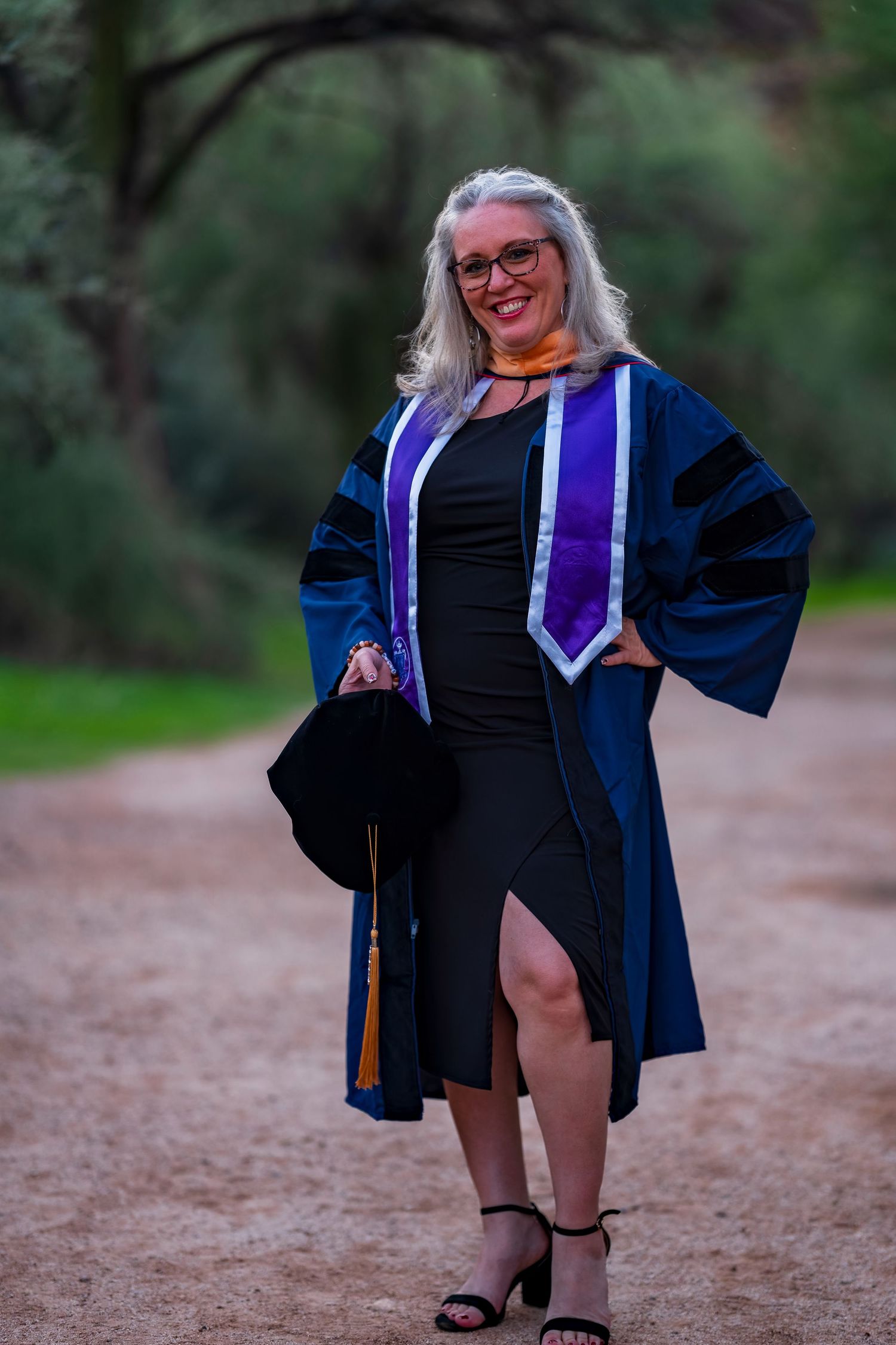 Graduate in blue doctoral robes and black dress poses on dirt path with trees in background.