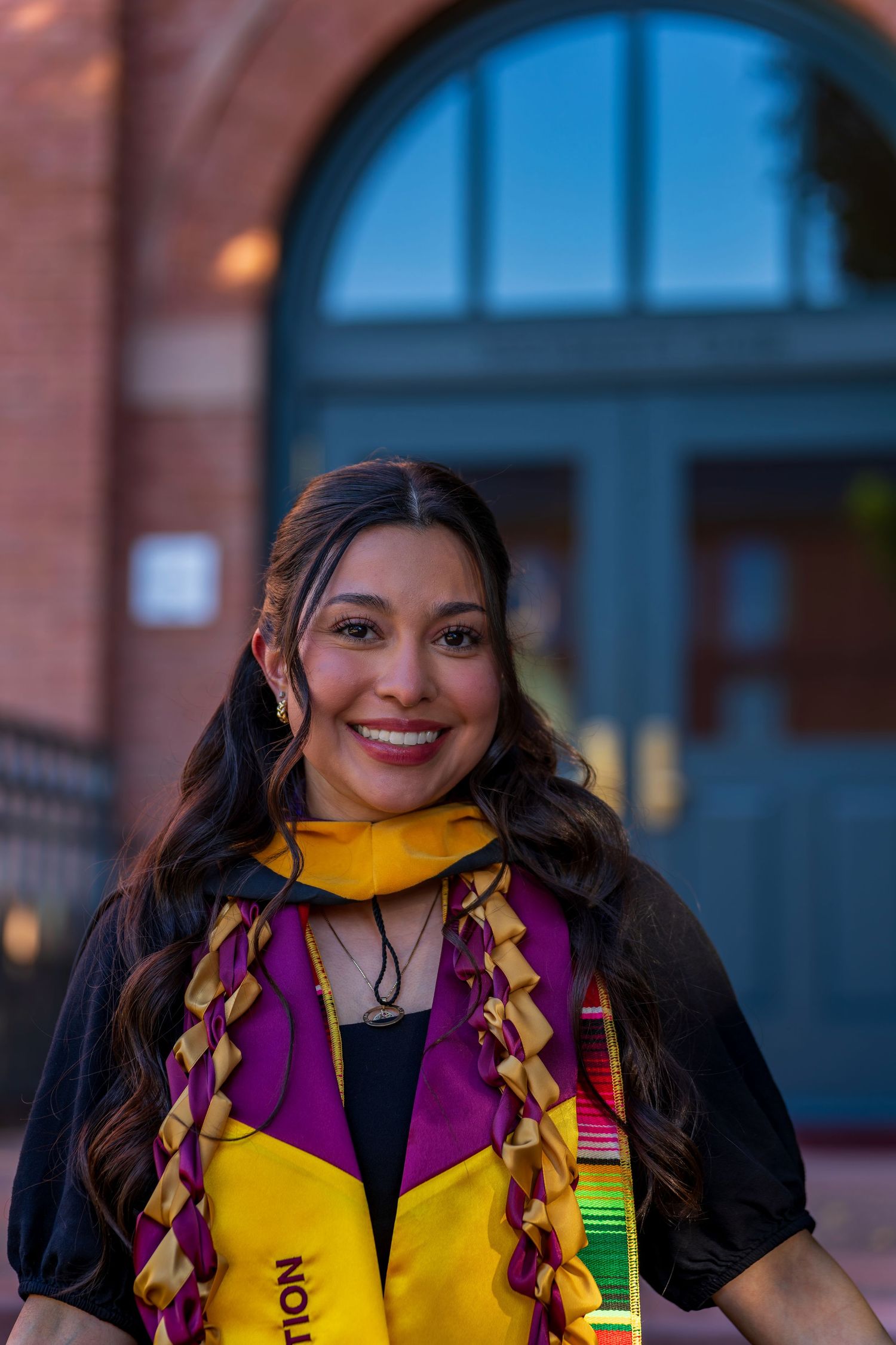 Graduate in black gown with colorful stole smiling in front of brick building with blue doors.