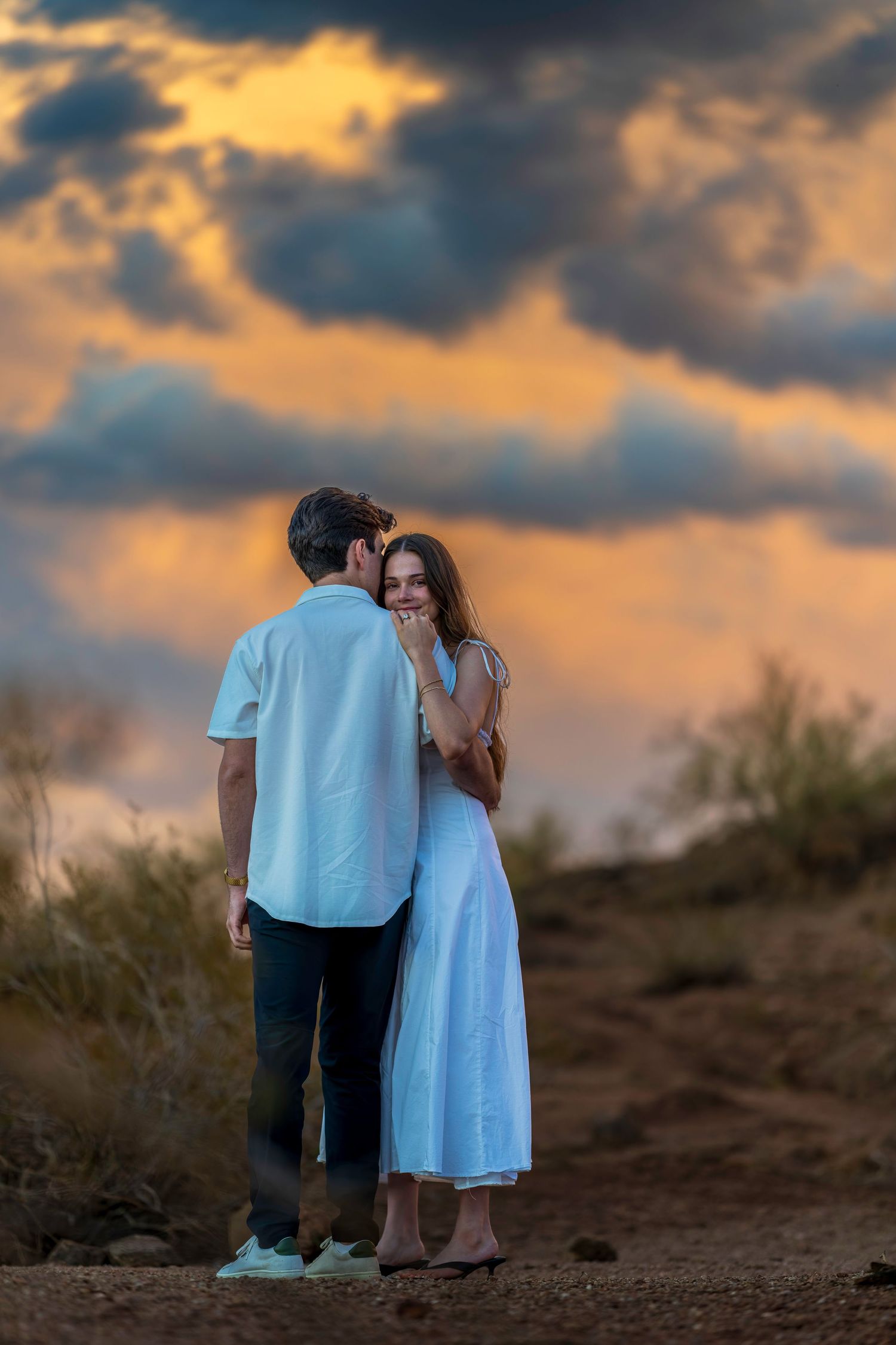 A couple stands together on a hilltop against a dramatic orange and blue sunset sky.