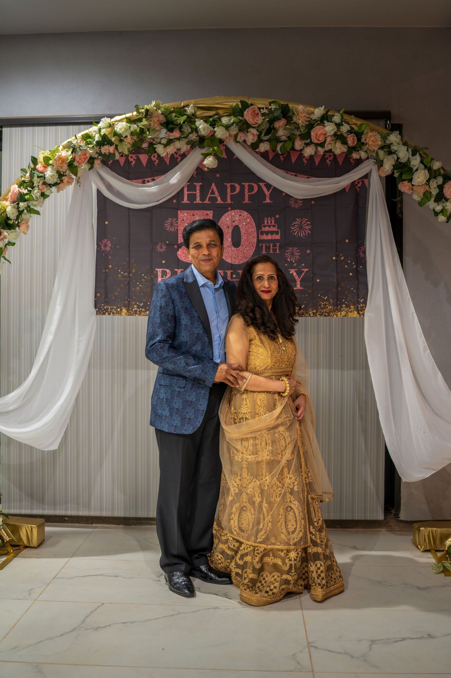 Decorated photo backdrop with white drapes, floral arch and gold 50th birthday signage.