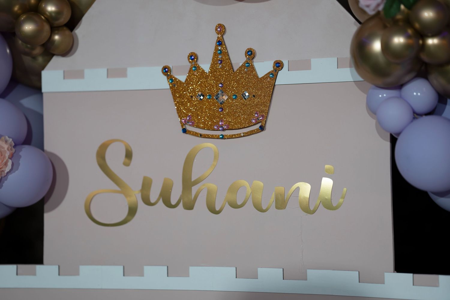 Gold glittery crown decoration and text reading 'Suhani' against a backdrop of purple and gold balloons.