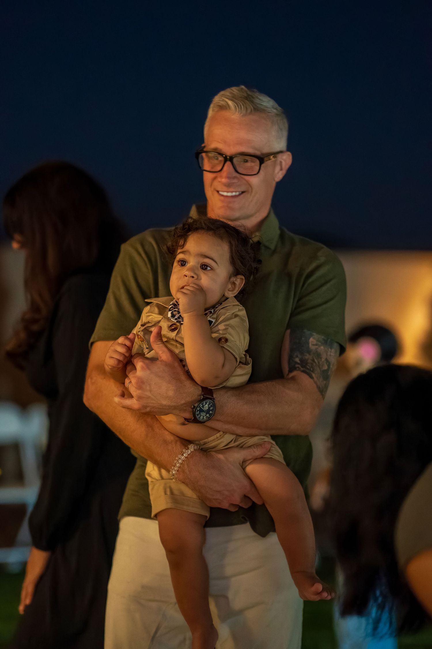 Adult in glasses and olive shirt holds young child at evening outdoor gathering.