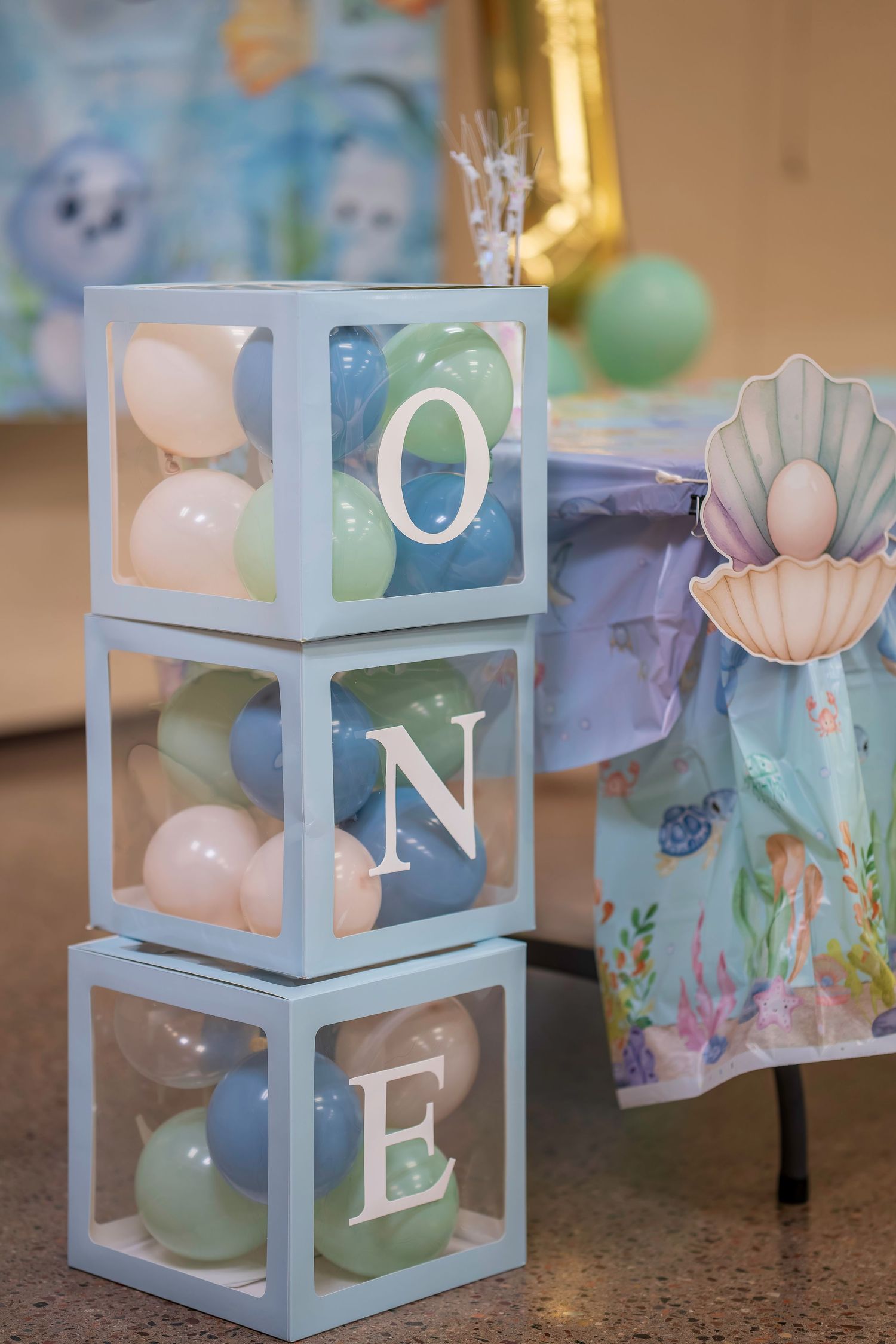 Three clear boxes stacked spelling ONE filled with pastel balloons at an ocean-themed first birthday party.