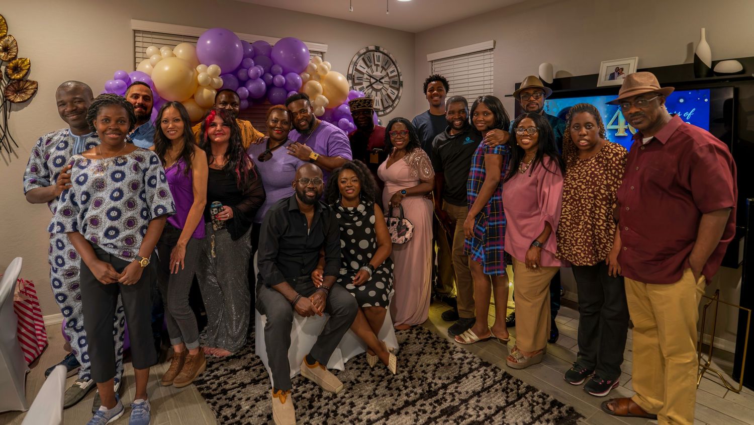 Large group of Black family and friends posing together at a celebration with purple and gold balloon decorations.