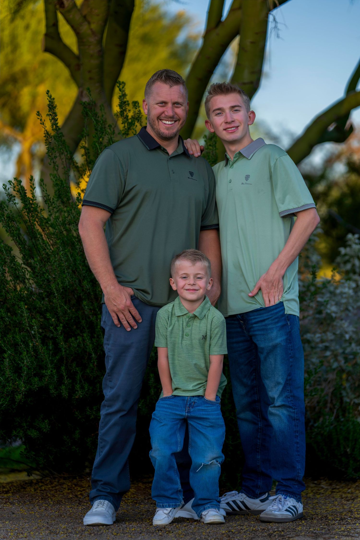 A smiling man and two boys wearing matching green polos pose together outdoors near trees at golden hour.