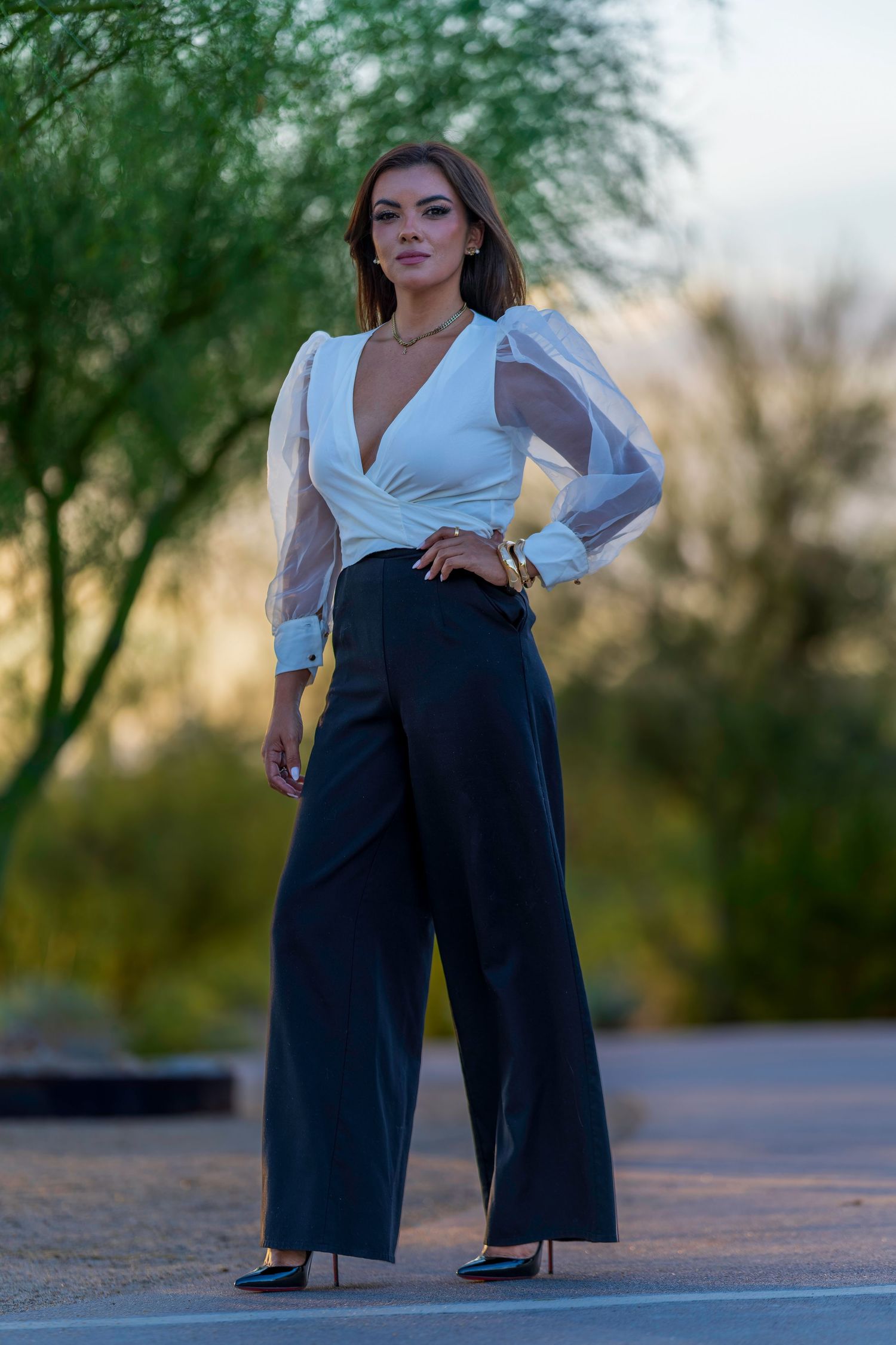 Elegant woman in white sheer blouse and black wide-leg pants posing outdoors at sunset.