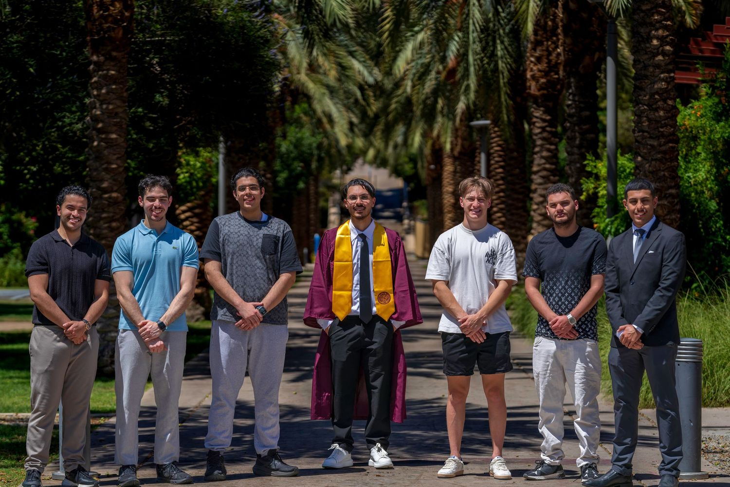 Group of seven young men posing outdoors at a graduation celebration surrounded by palm trees.