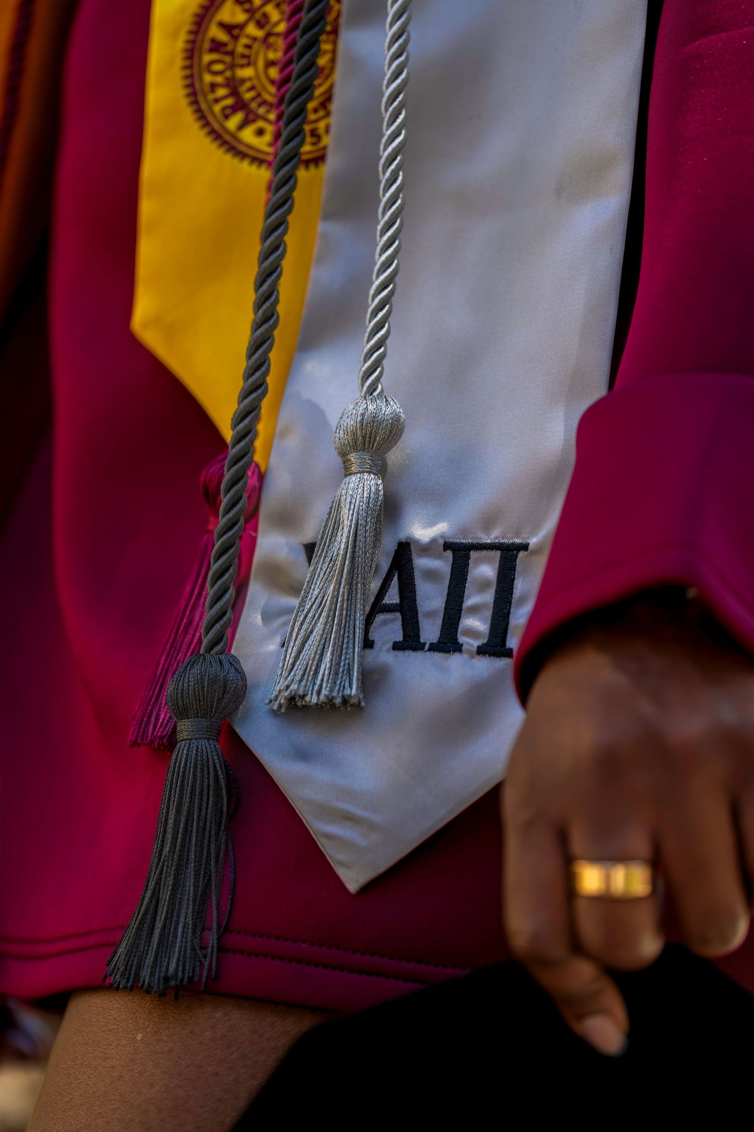 Graduate wearing maroon gown with white stole, honor cords, and gold ring at college commencement ceremony.
