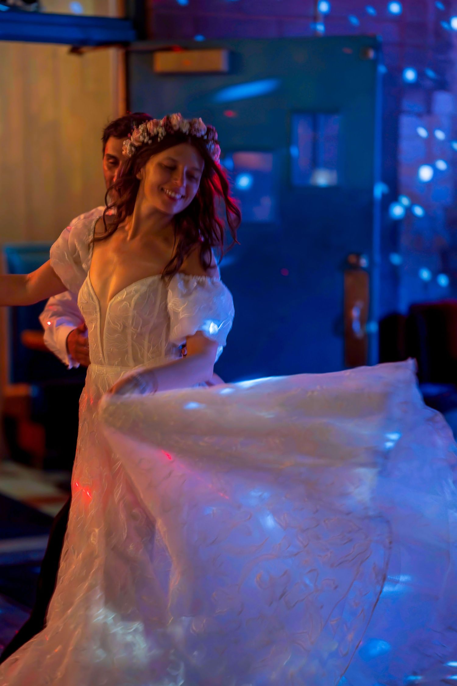 Bride in white gown with floral crown dancing with groom under colorful disco lights at wedding reception.