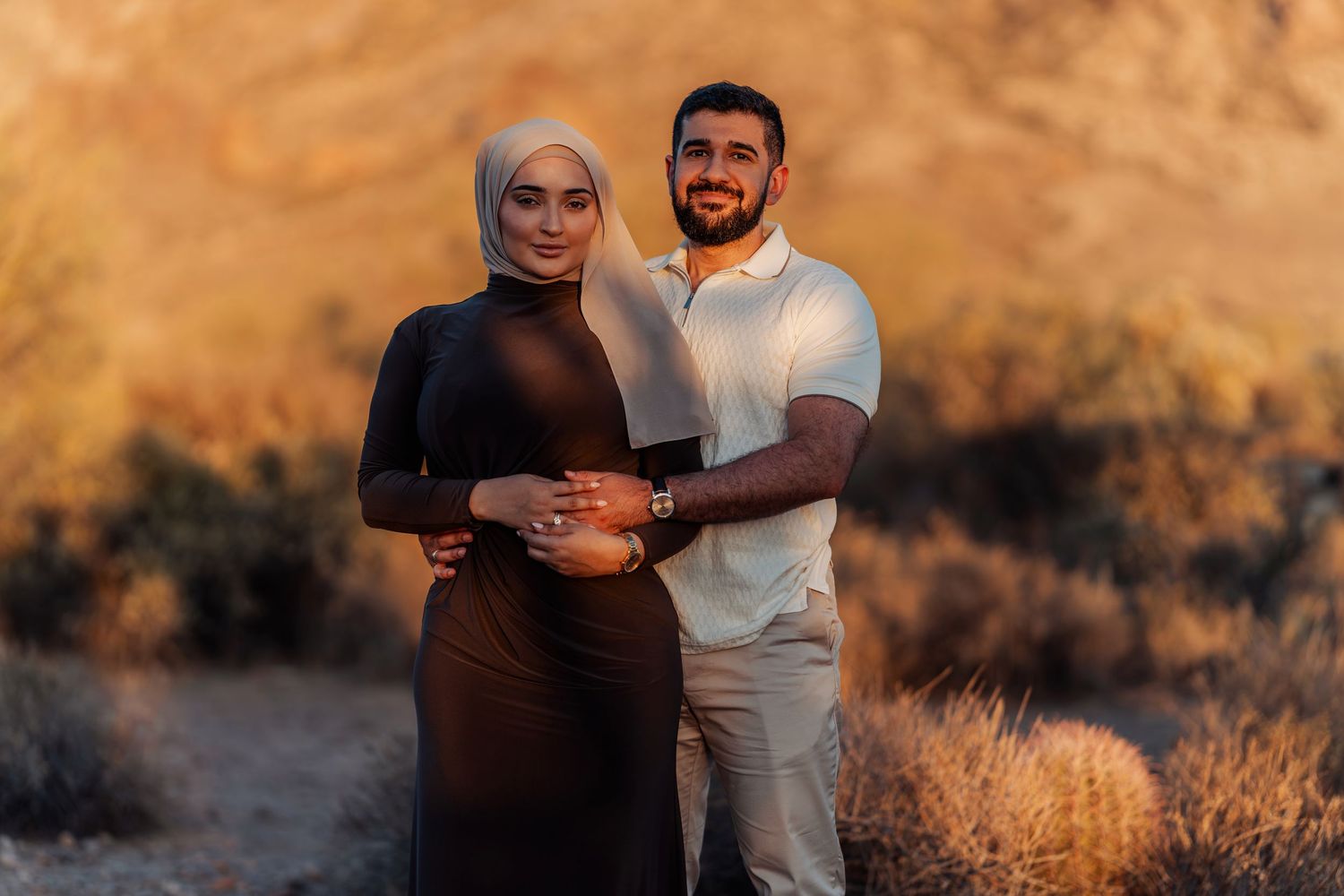 A couple poses together at golden hour in a desert landscape, woman wearing hijab and brown dress.