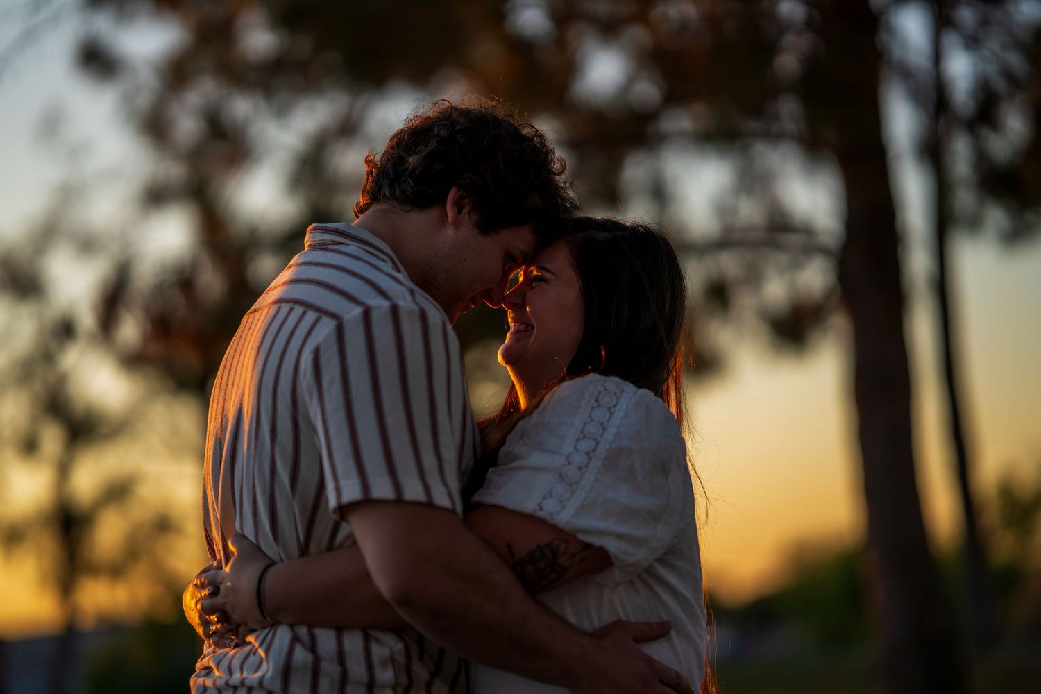 Couple sharing a tender almost-kiss moment at sunset surrounded by blurred trees in warm golden light.