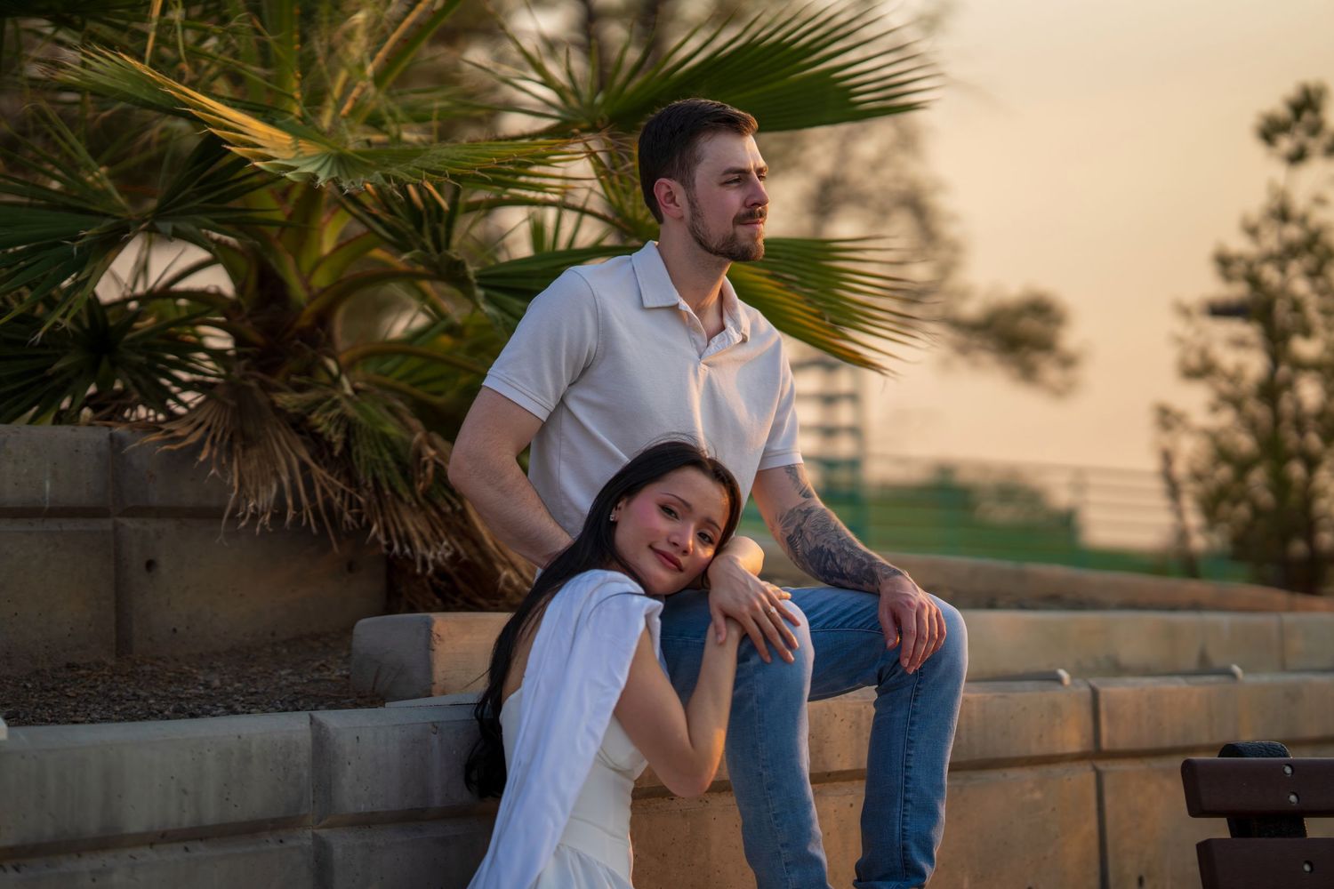 Couple relaxing at sunset near a palm tree, woman leans on man seated on stone wall.