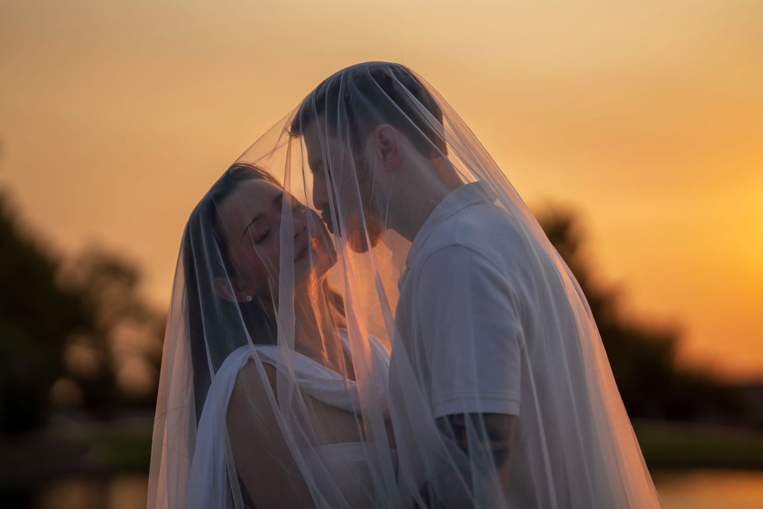 Newlyweds sharing a kiss under a flowing veil against a vibrant orange sunset sky.