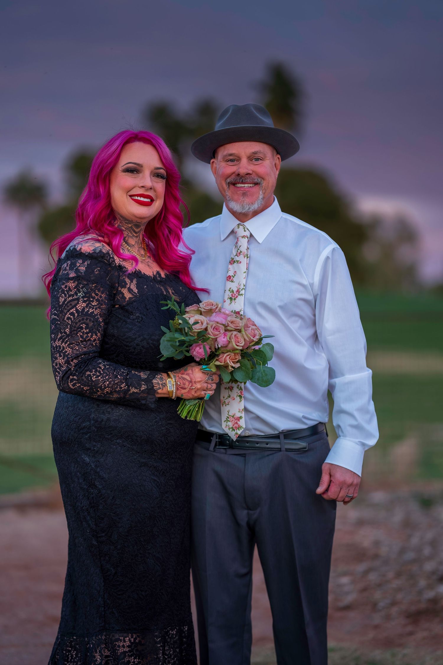 Couple posing outdoors at sunset, woman with pink hair in black lace dress holding bouquet, man in white shirt and gray hat.