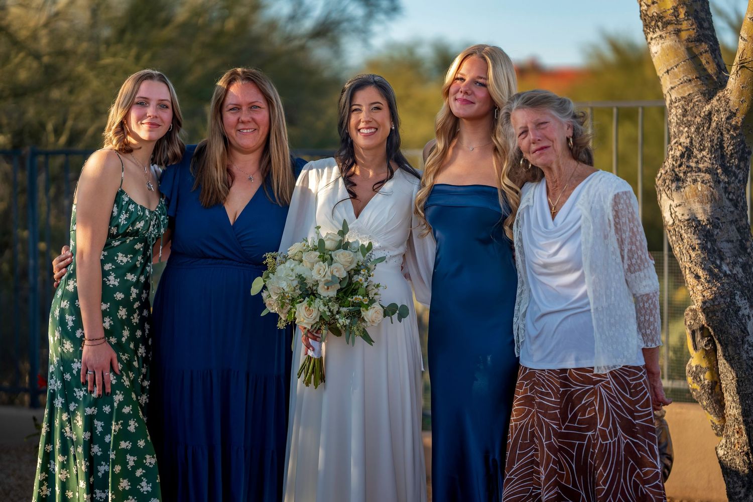 Bride in white gown poses with four women in blue and green dresses outdoors at wedding.