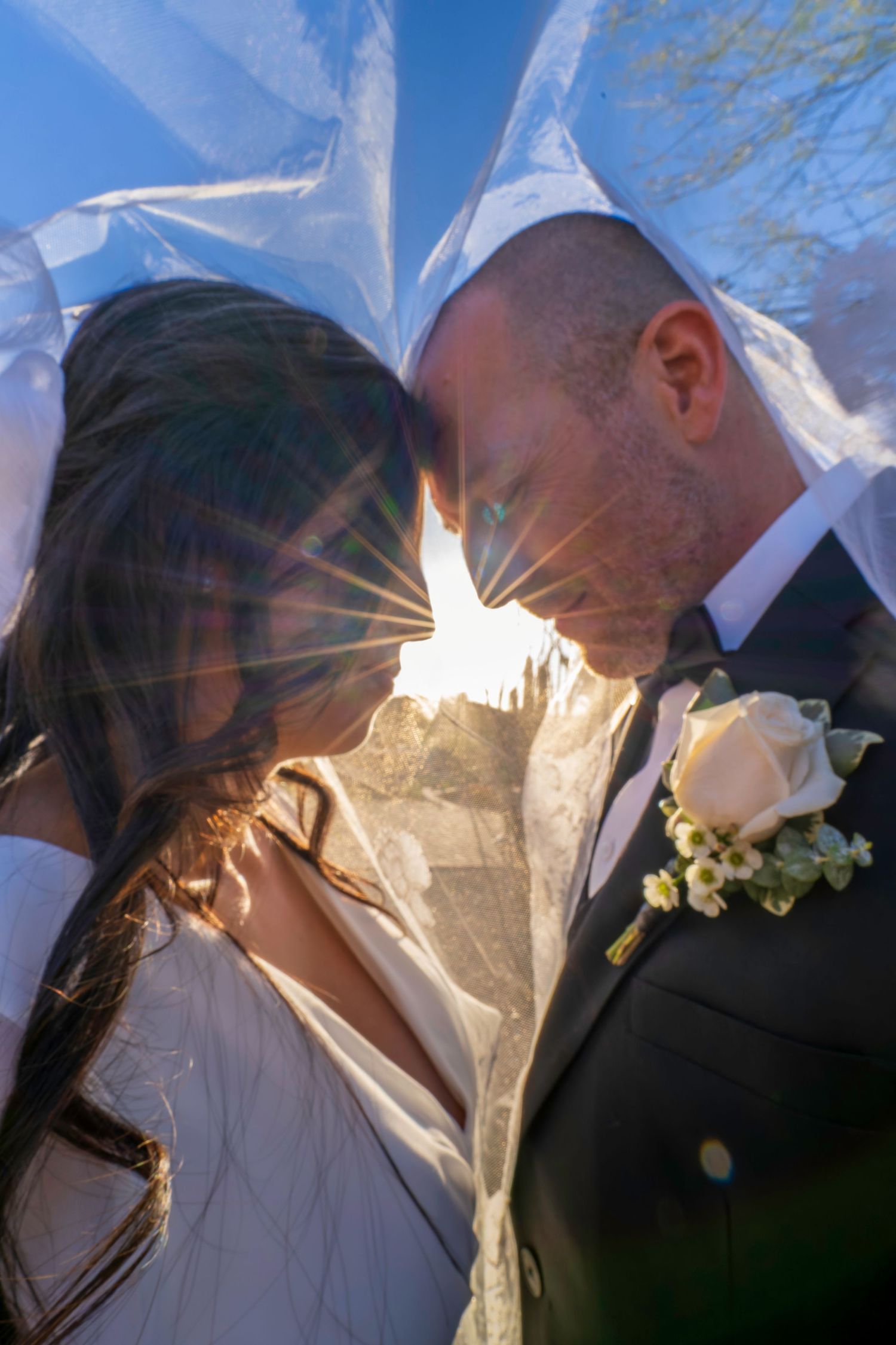 Bride and groom touching foreheads under sheer veil with stunning sunburst light effect between them.