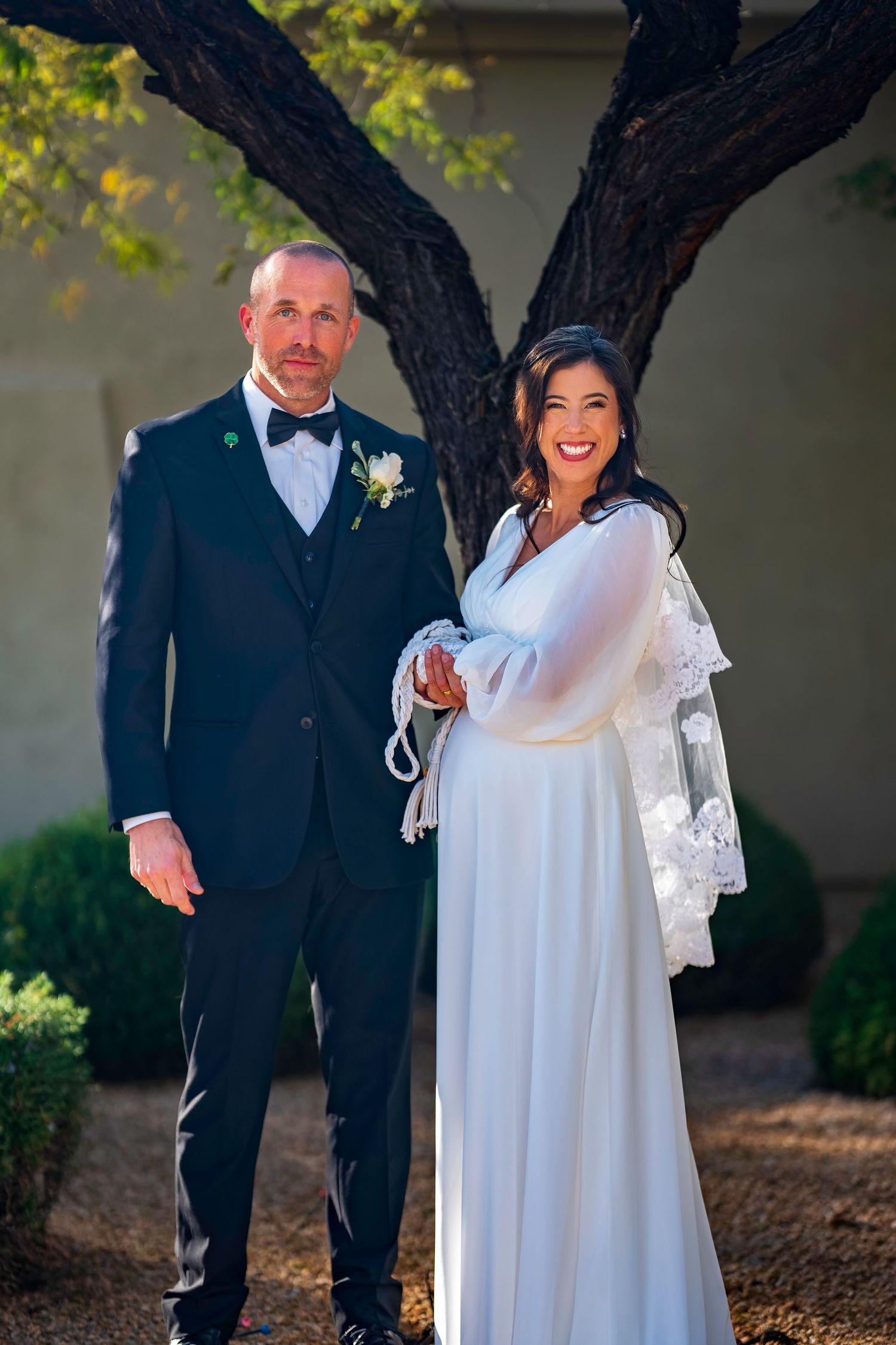 Smiling bride and groom posing together under a tree in outdoor wedding portrait.