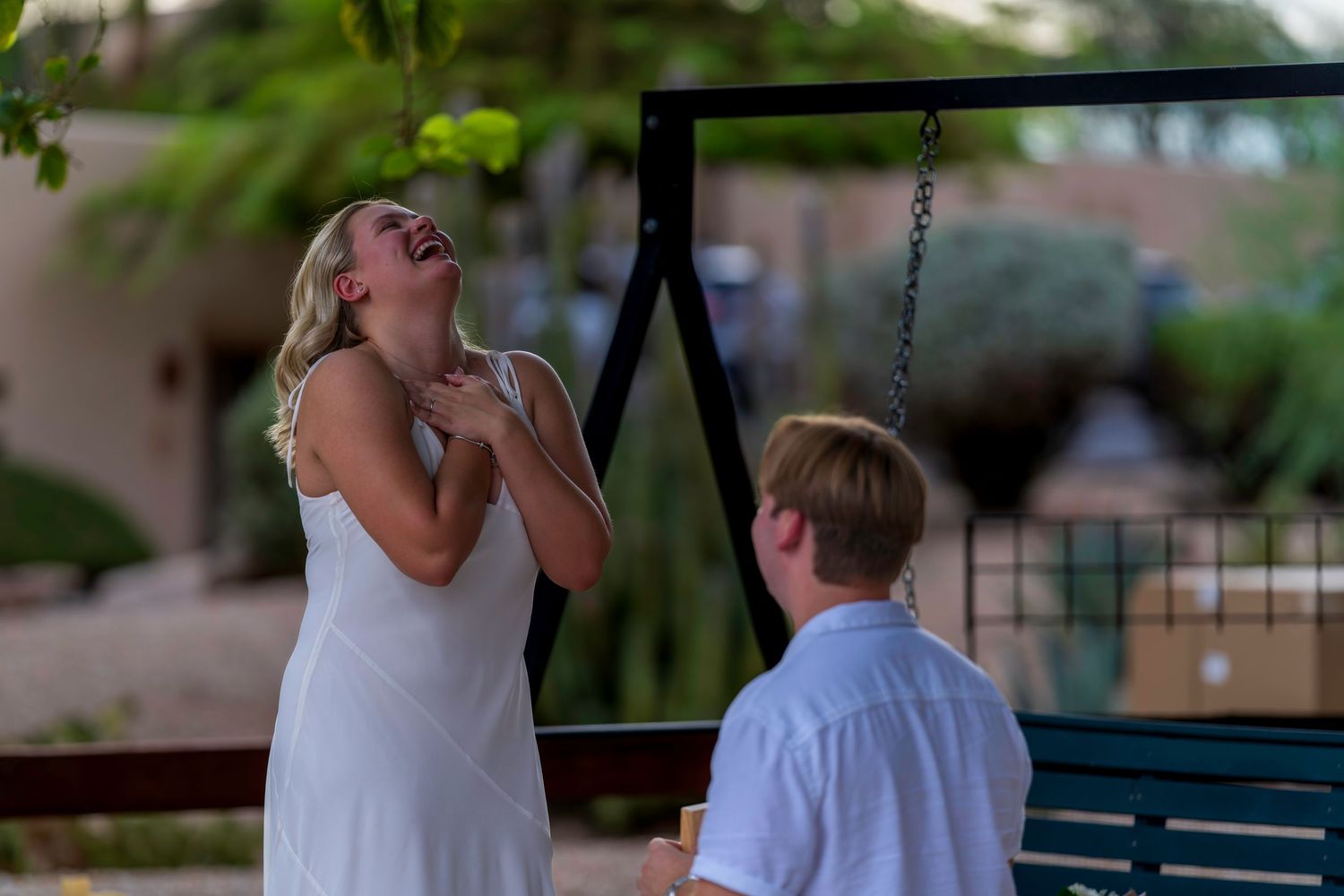 Woman in white dress laughing joyfully during an outdoor marriage proposal near a swing set.