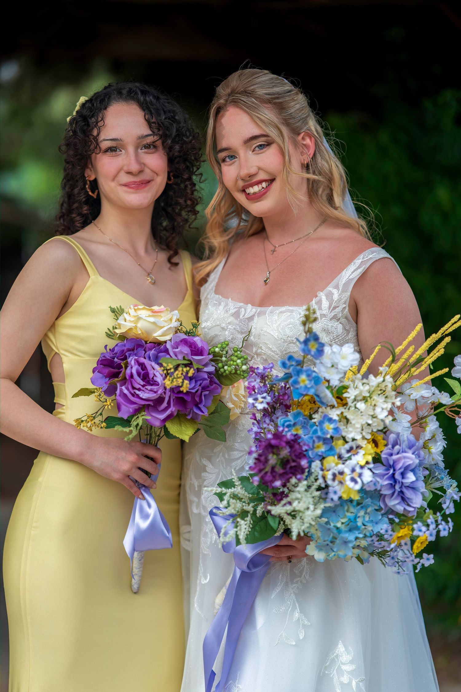 Bride in white gown and bridesmaid in yellow dress holding colorful purple and blue flower bouquets at wedding.