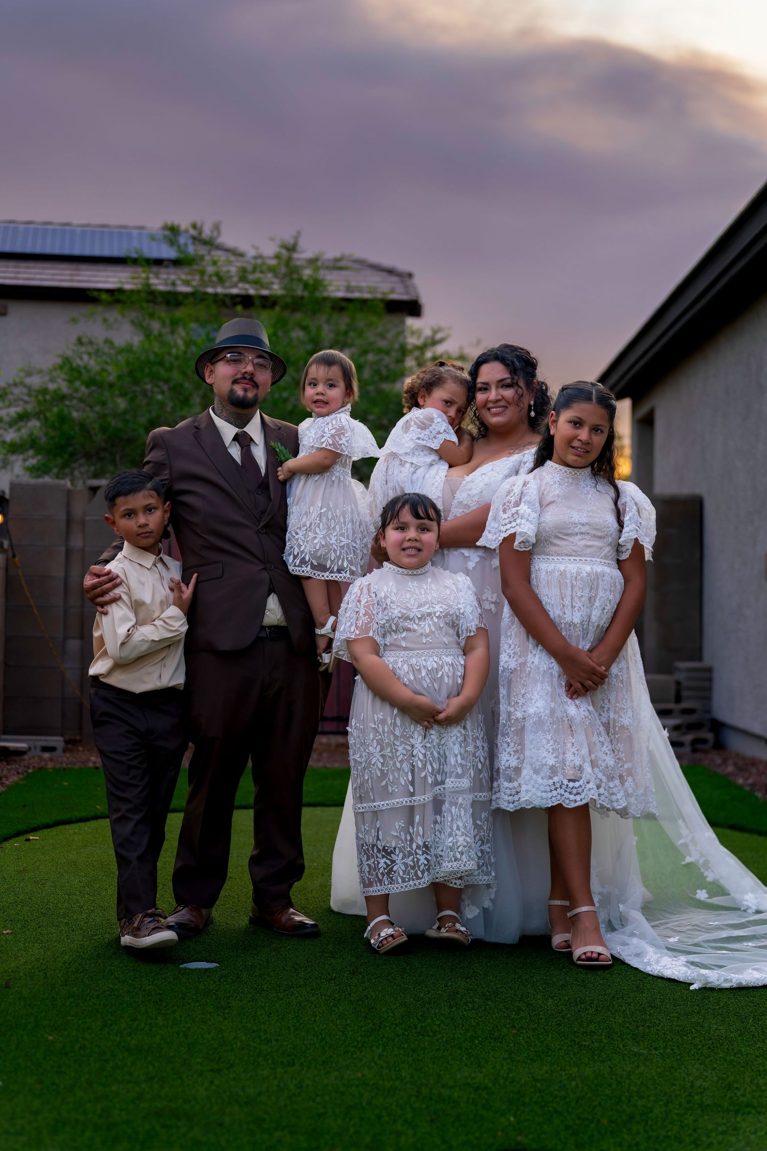 Family portrait at outdoor wedding with bride, groom, and children in white lace dresses and suits at dusk.
