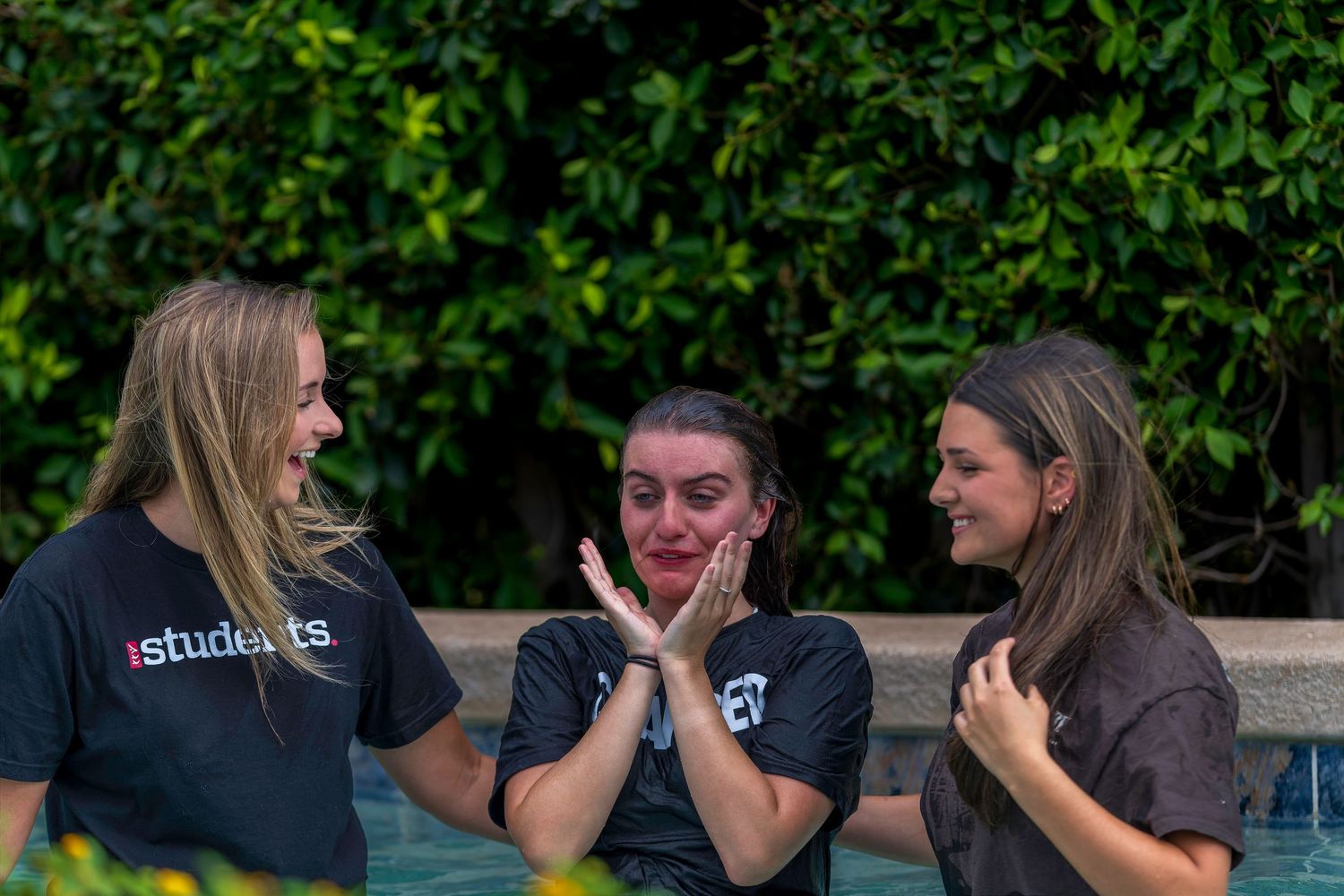Three young women standing by a pool, one emotional with hands raised, wearing matching black students shirts.
