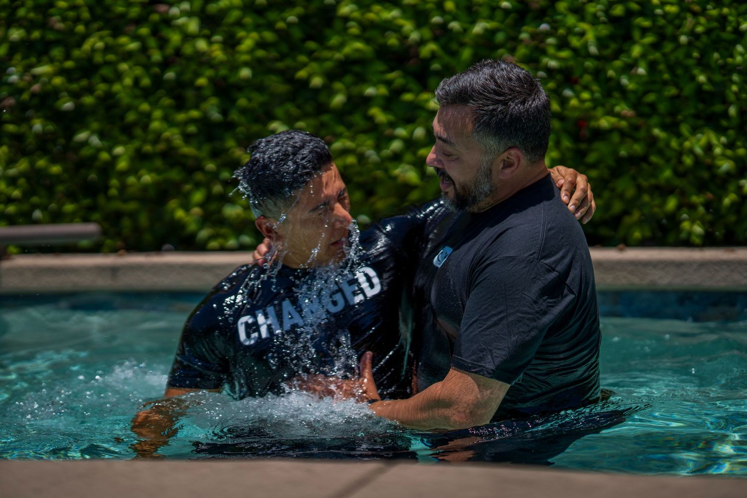 Man in a 'Changed' shirt being baptized in a pool by another man on a sunny day.