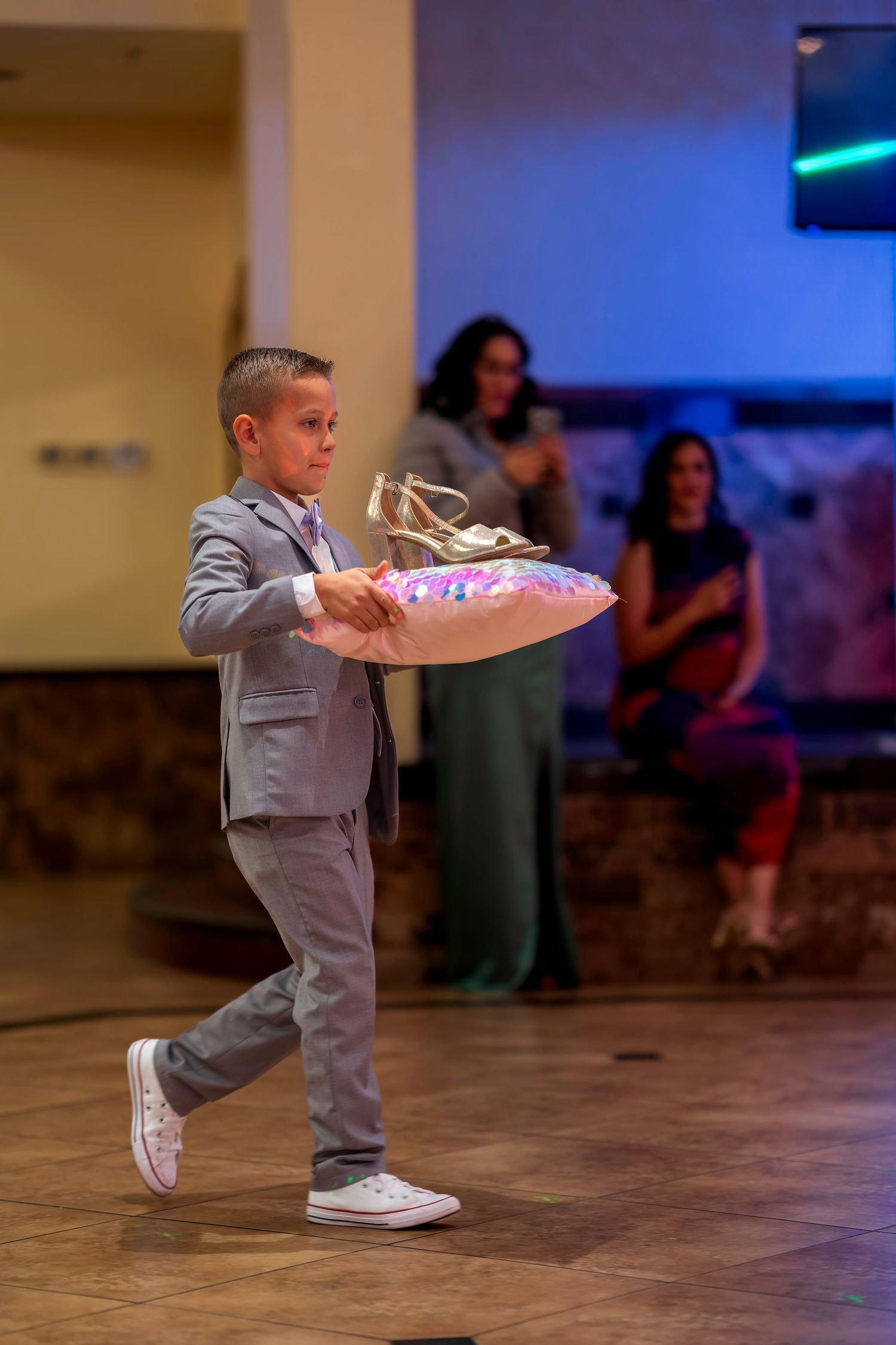 Young boy in gray suit carrying illuminated pillow with shoes at formal event ceremony.