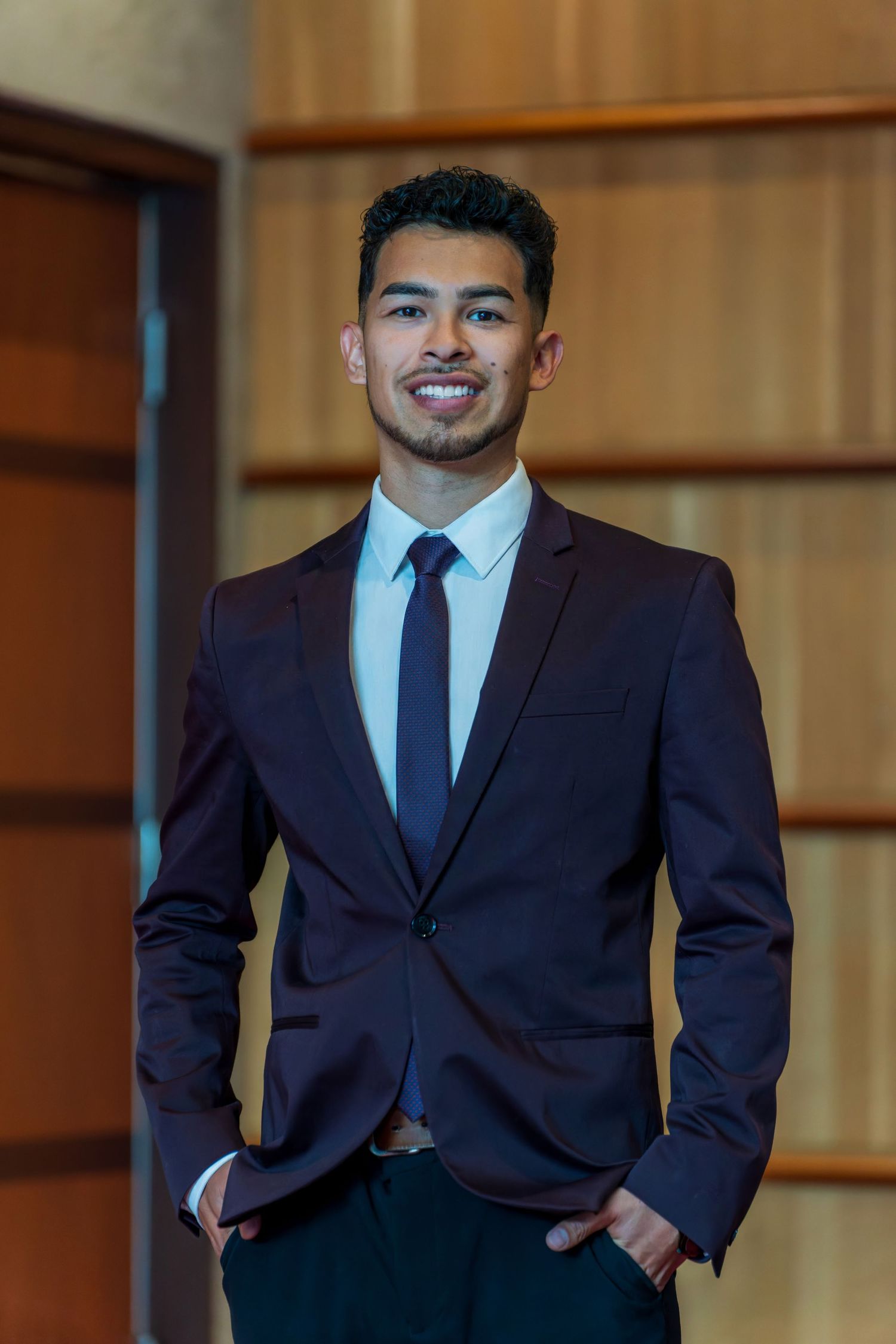 Professional indoor portrait of person in navy suit against wooden paneled wall.