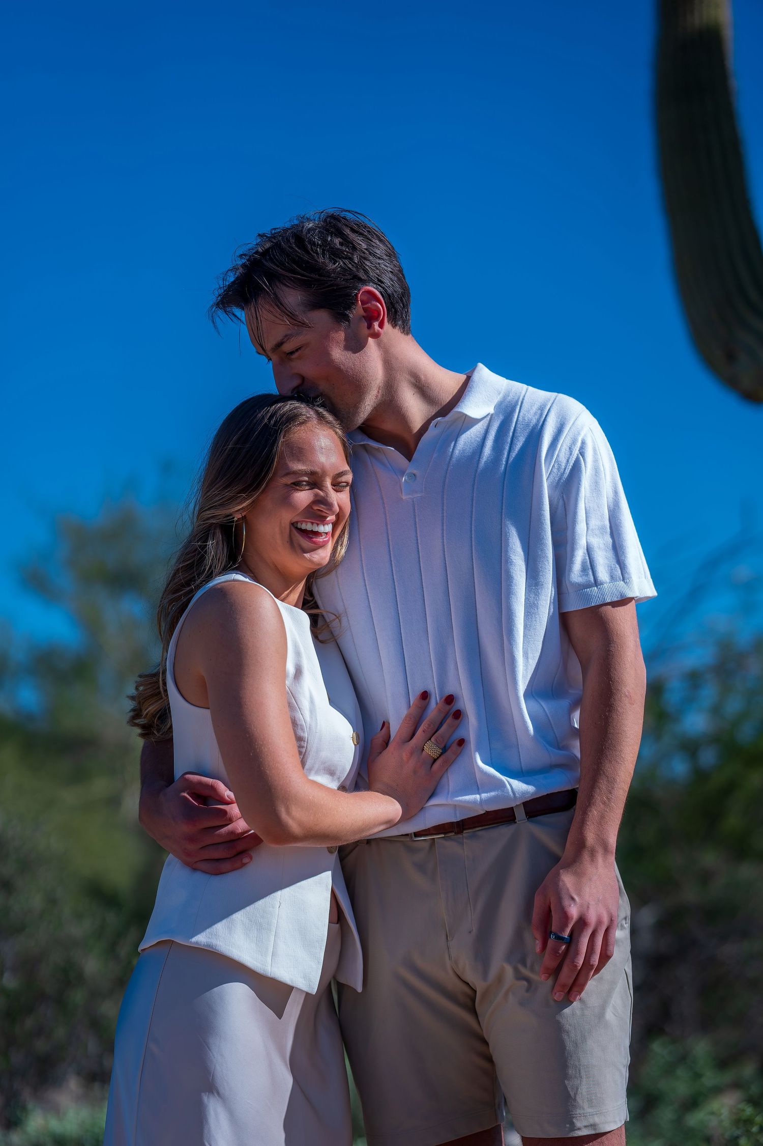 Romantic desert photoshoot featuring couple in white tops against cacti and blue sky.