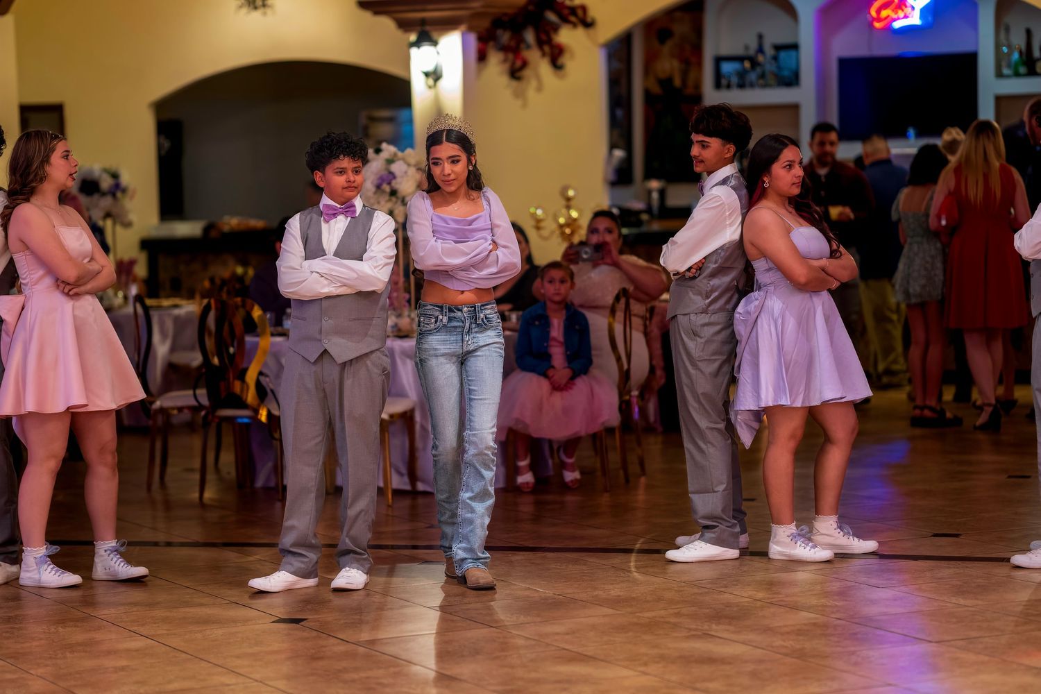 Young people in formal attire stand on a dance floor at an elegant indoor event or school dance.