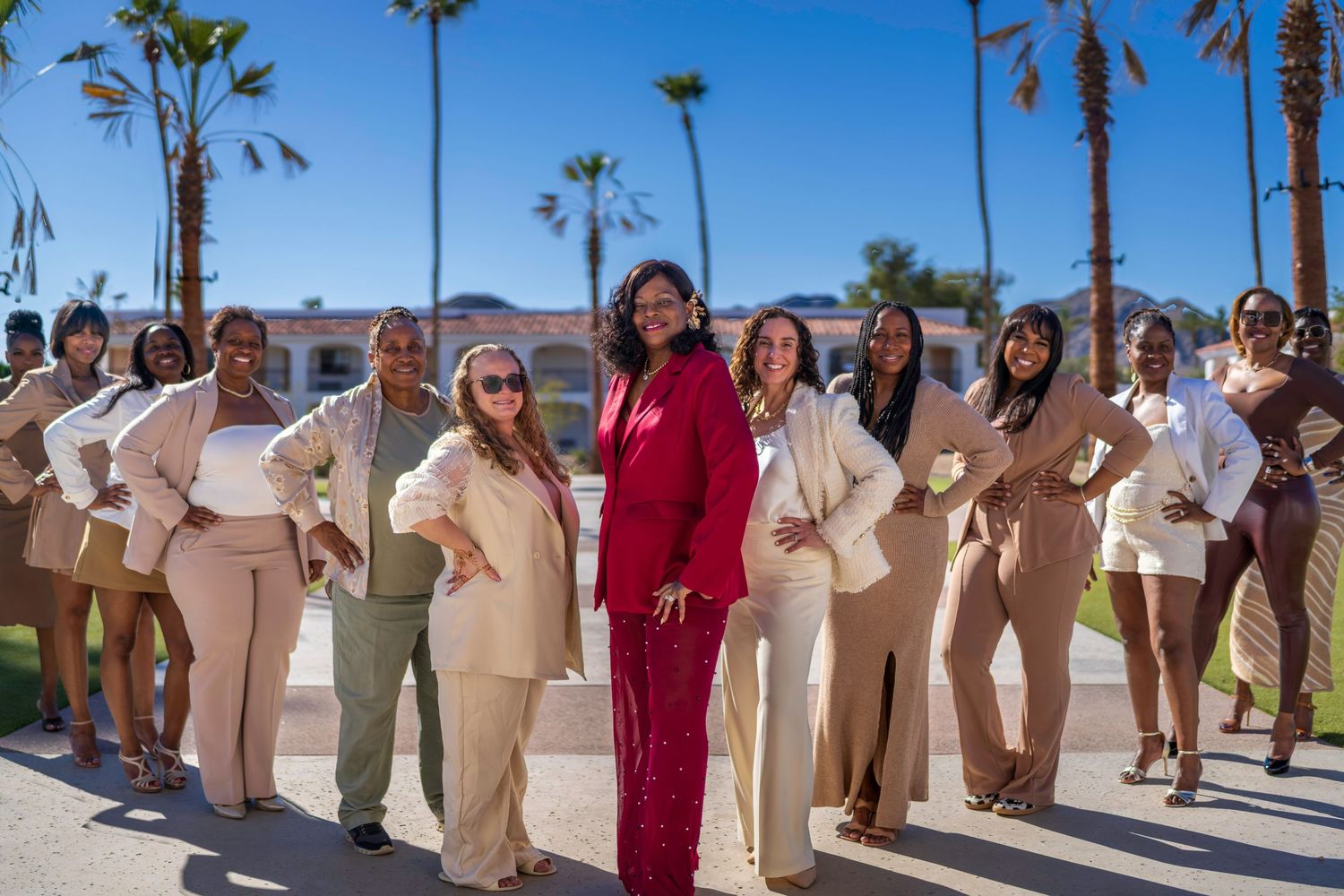 Group of women in neutral outfits posing outdoors with palm trees and blue sky backdrop.