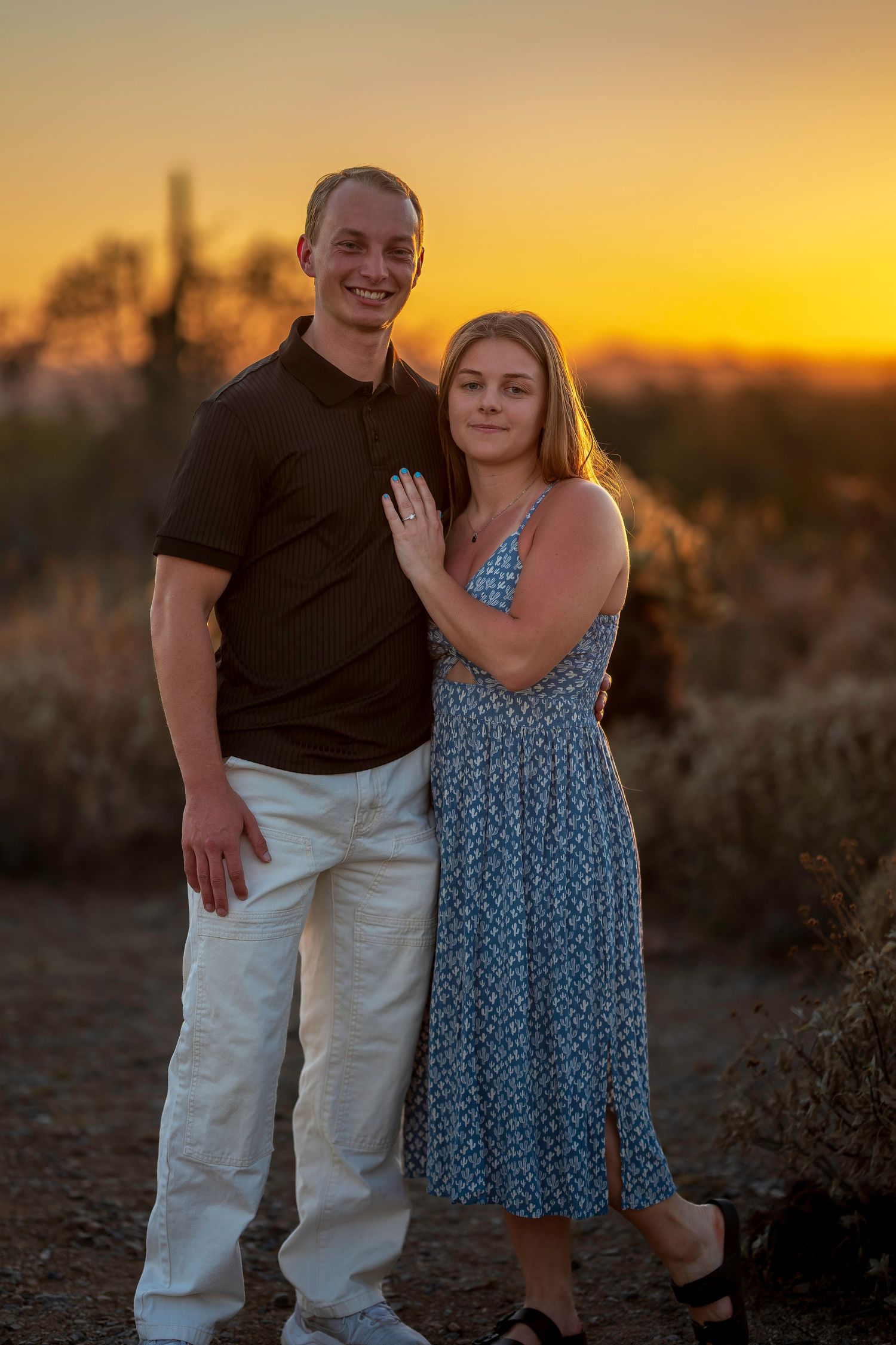 Couple posing together at golden hour sunset in Arizona desert, woman in blue floral dress, man in black polo.