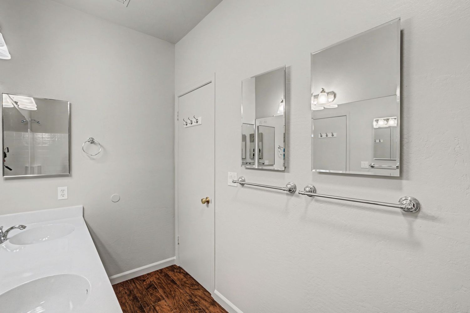 Bright white bathroom with dual sinks, large mirrors, grab bars, and dark hardwood floors.