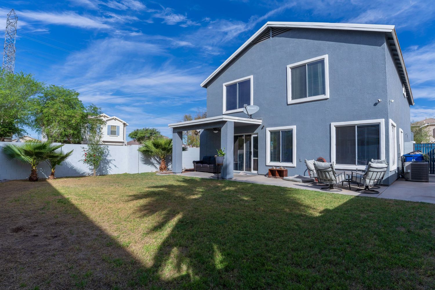 Two-story gray stucco home with spacious green backyard, patio seating area, and palm tree under blue sky.