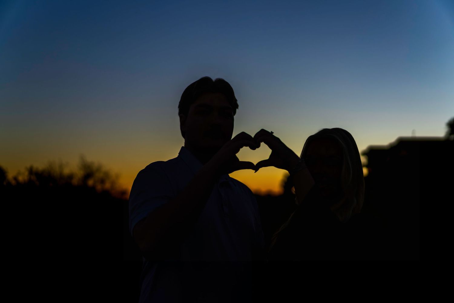 Couple silhouetted against a vibrant sunset making a heart shape with their hands.