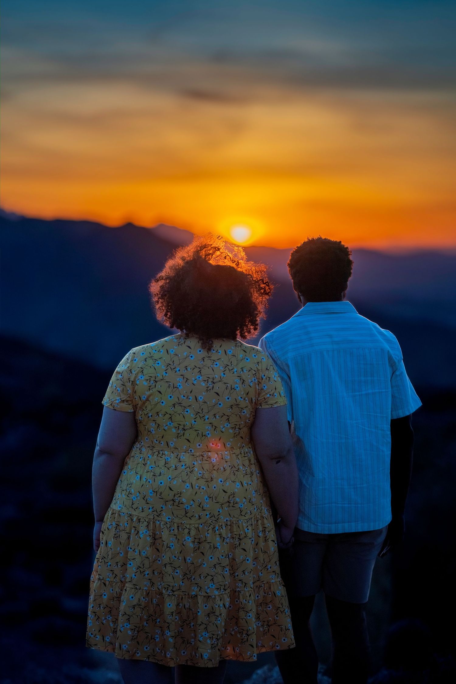 Romantic couple holding hands watching a vibrant orange sunset over mountain silhouettes from behind.