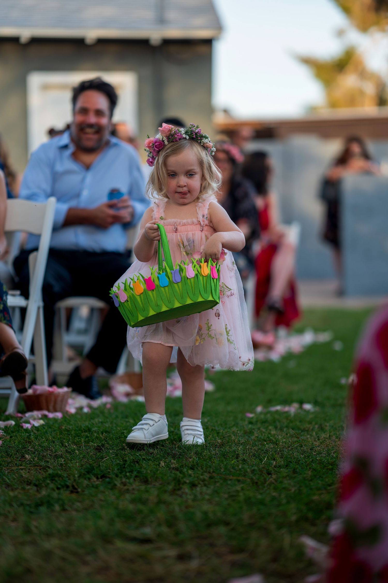 Adorable flower girl in pink dress carrying colorful basket down outdoor wedding aisle.