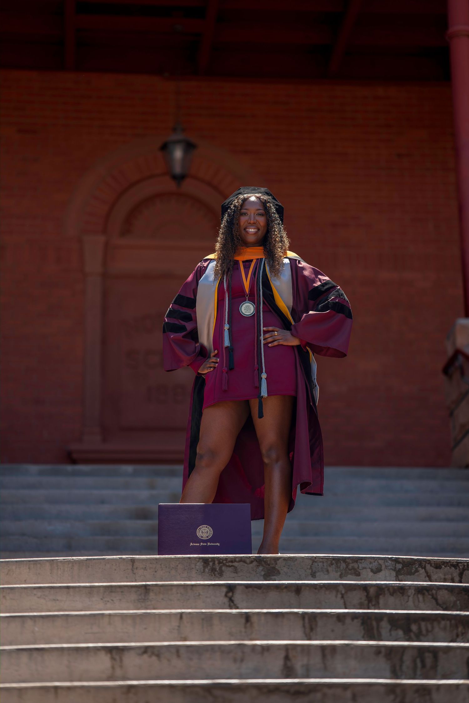 A smiling graduate in maroon regalia and medal stands proudly on steps outside a brick building holding a diploma.