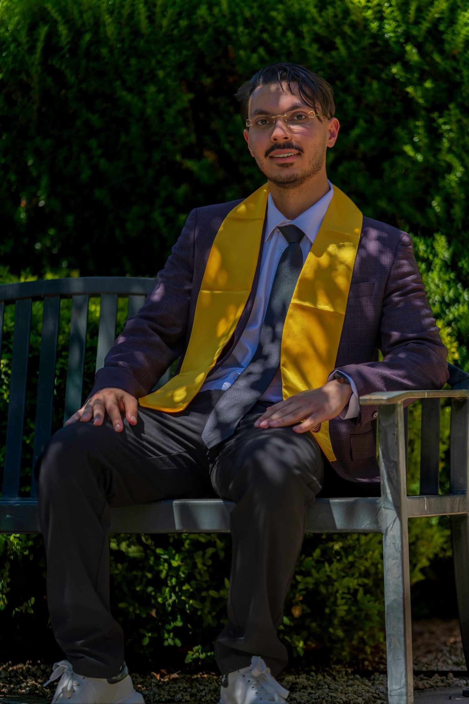 Graduate in suit and gold stole sitting on bench in garden.