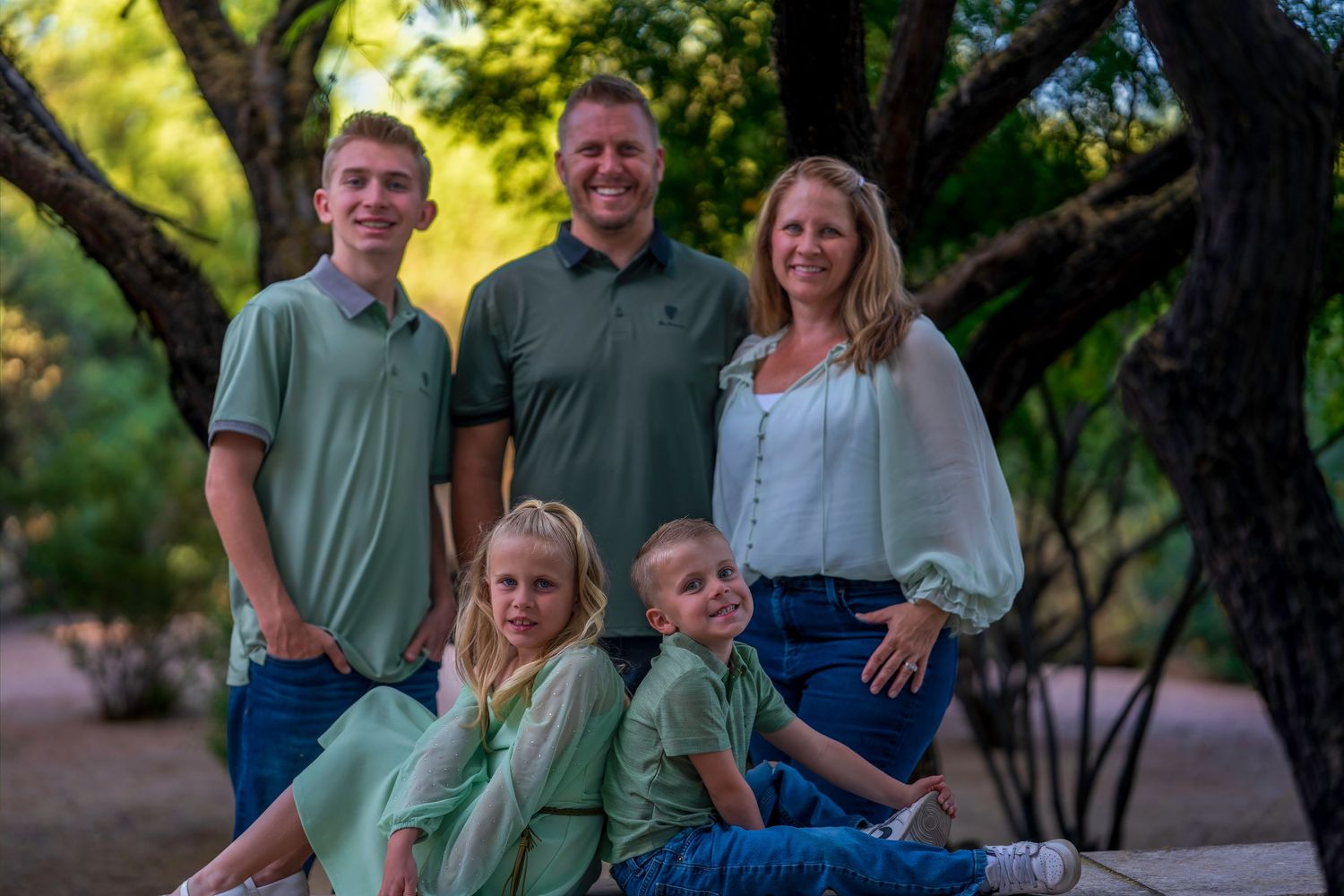 Family of five posing outdoors in coordinating green and white outfits among trees.