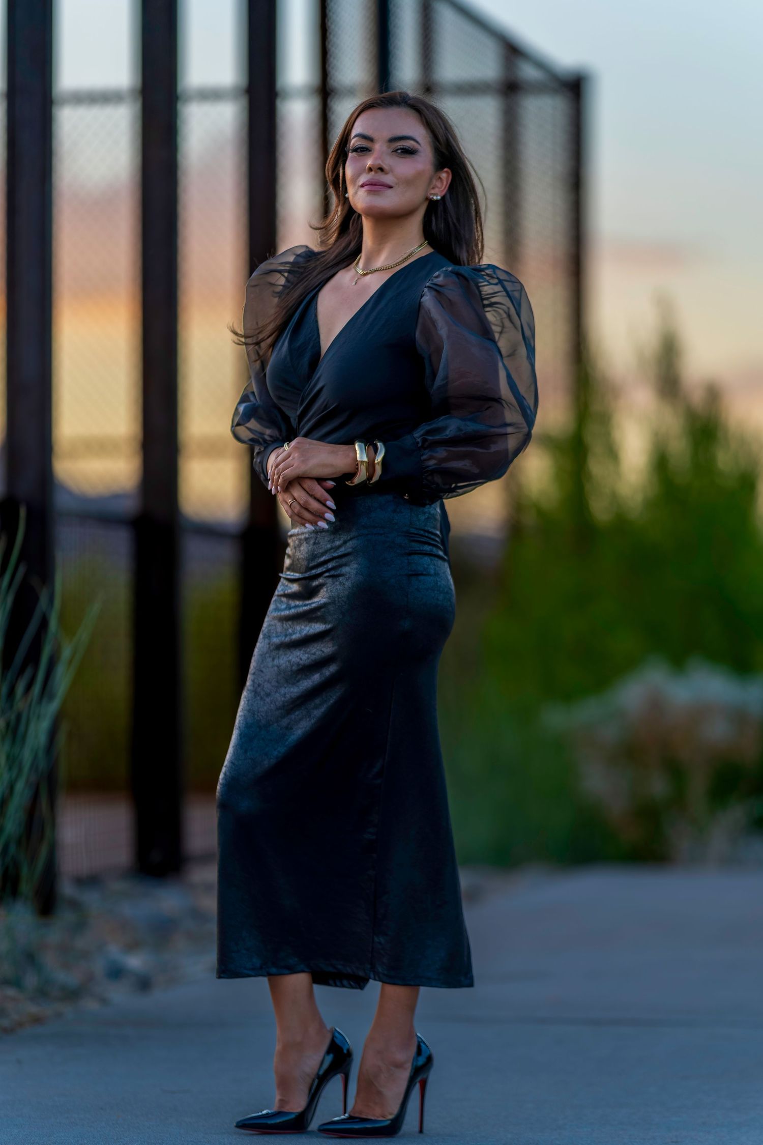 Elegant woman in black velvet midi dress and heels poses at sunset in outdoor urban setting.