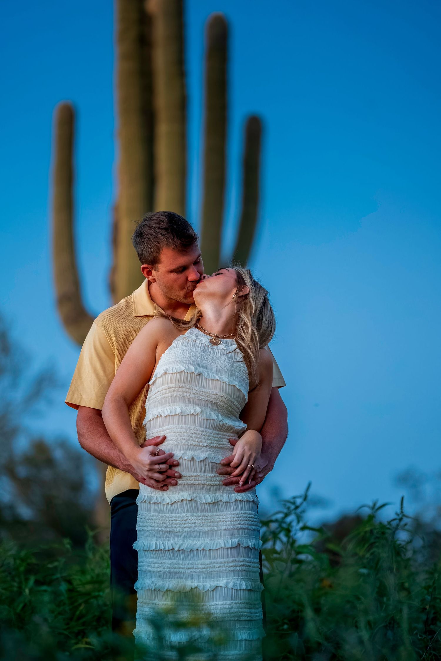 Couple kissing in front of tall saguaro cactus at dusk in Arizona desert landscape.