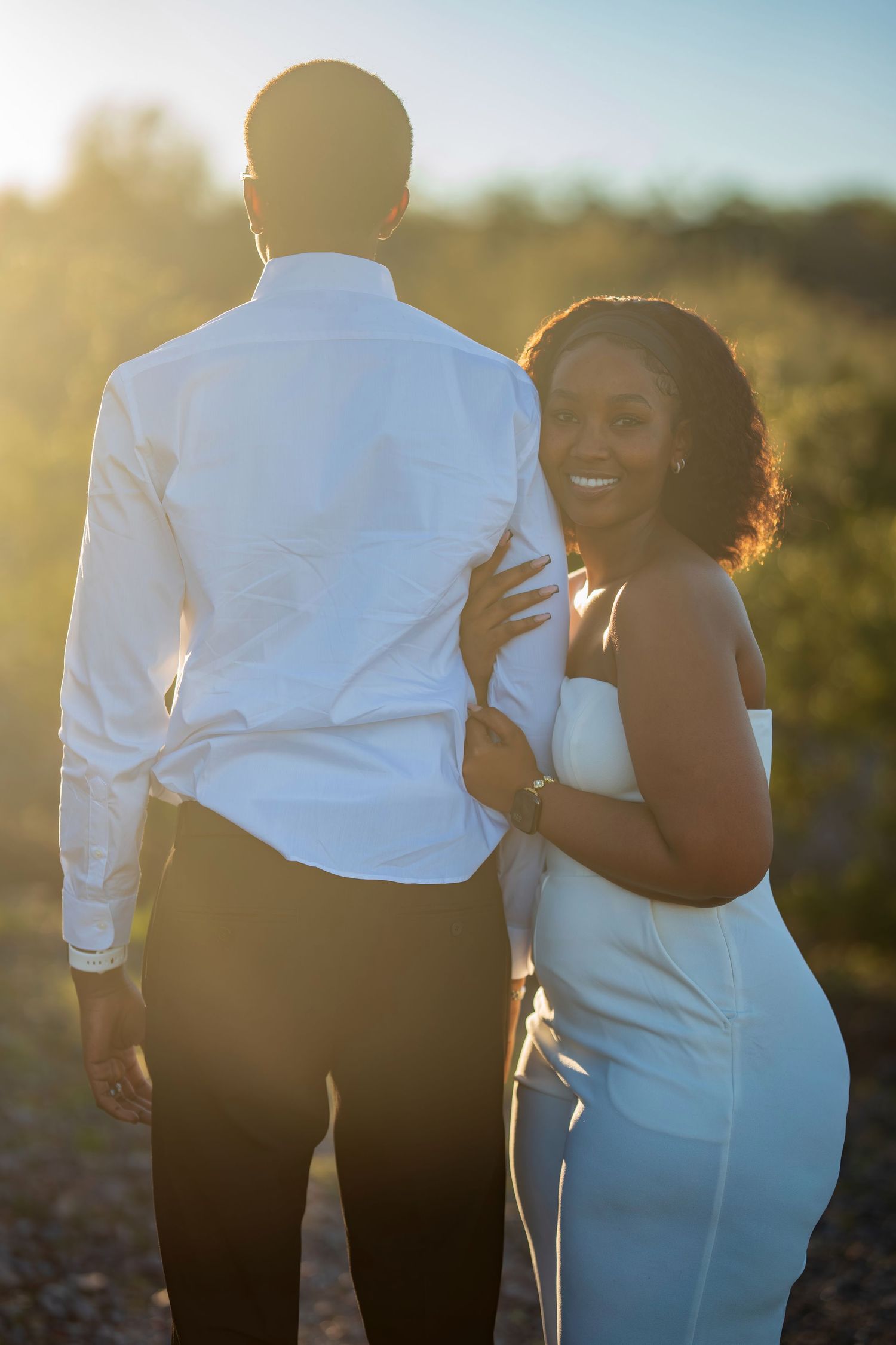 A smiling bride in white stands beside her groom backlit by golden sunset light.